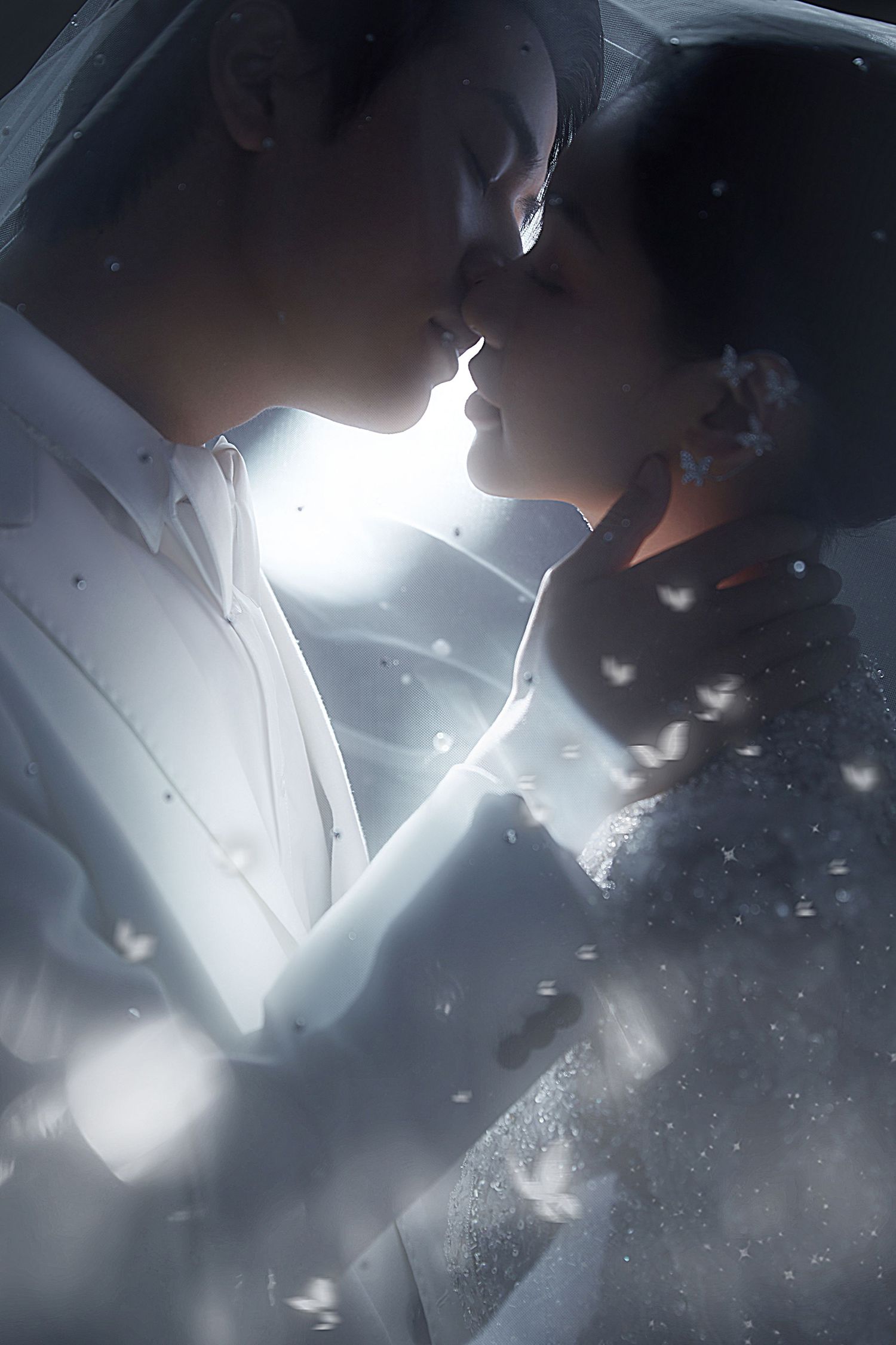 Wedding couple nearly kissing under veil with sparkling bokeh and dramatic backlit atmosphere.