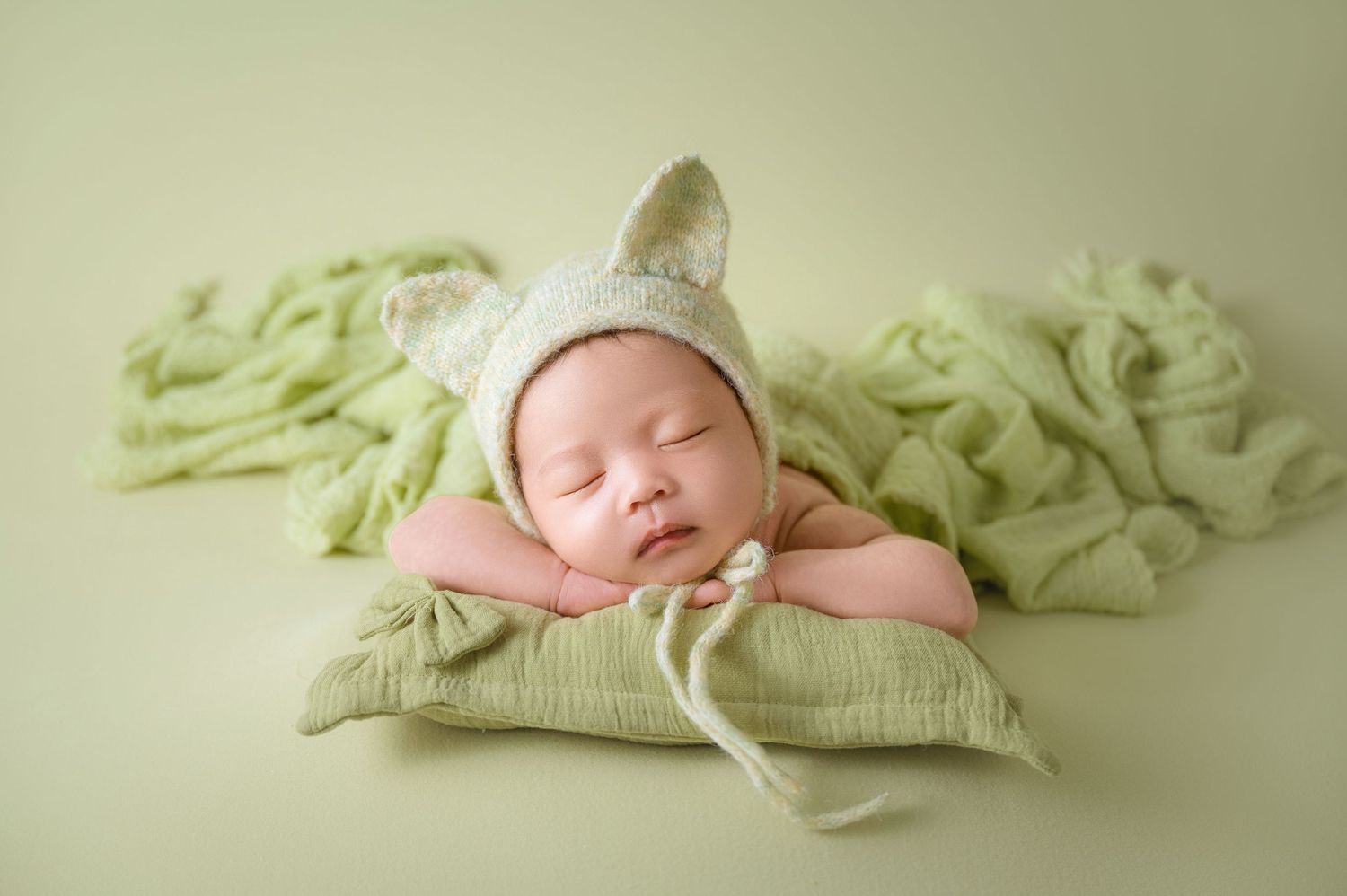 Sleeping newborn baby wearing a green bunny bonnet resting on a small pillow surrounded by soft green fabric.