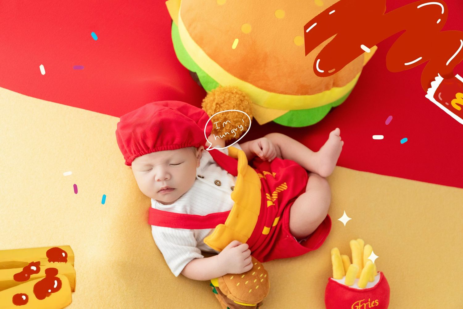 A sleeping baby dressed as a chef surrounded by fast food props including a burger, fries, and fried chicken on a colorful backdrop.