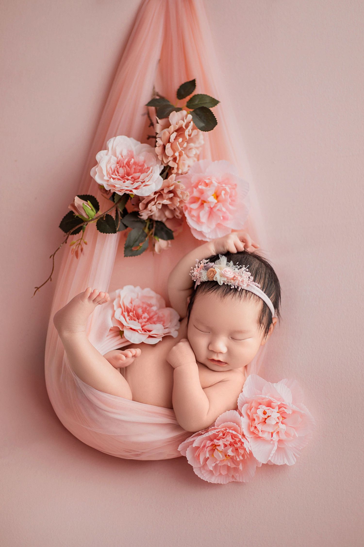 Sleeping newborn girl in pink floral headband nestled in blush tulle hammock with peony flowers.