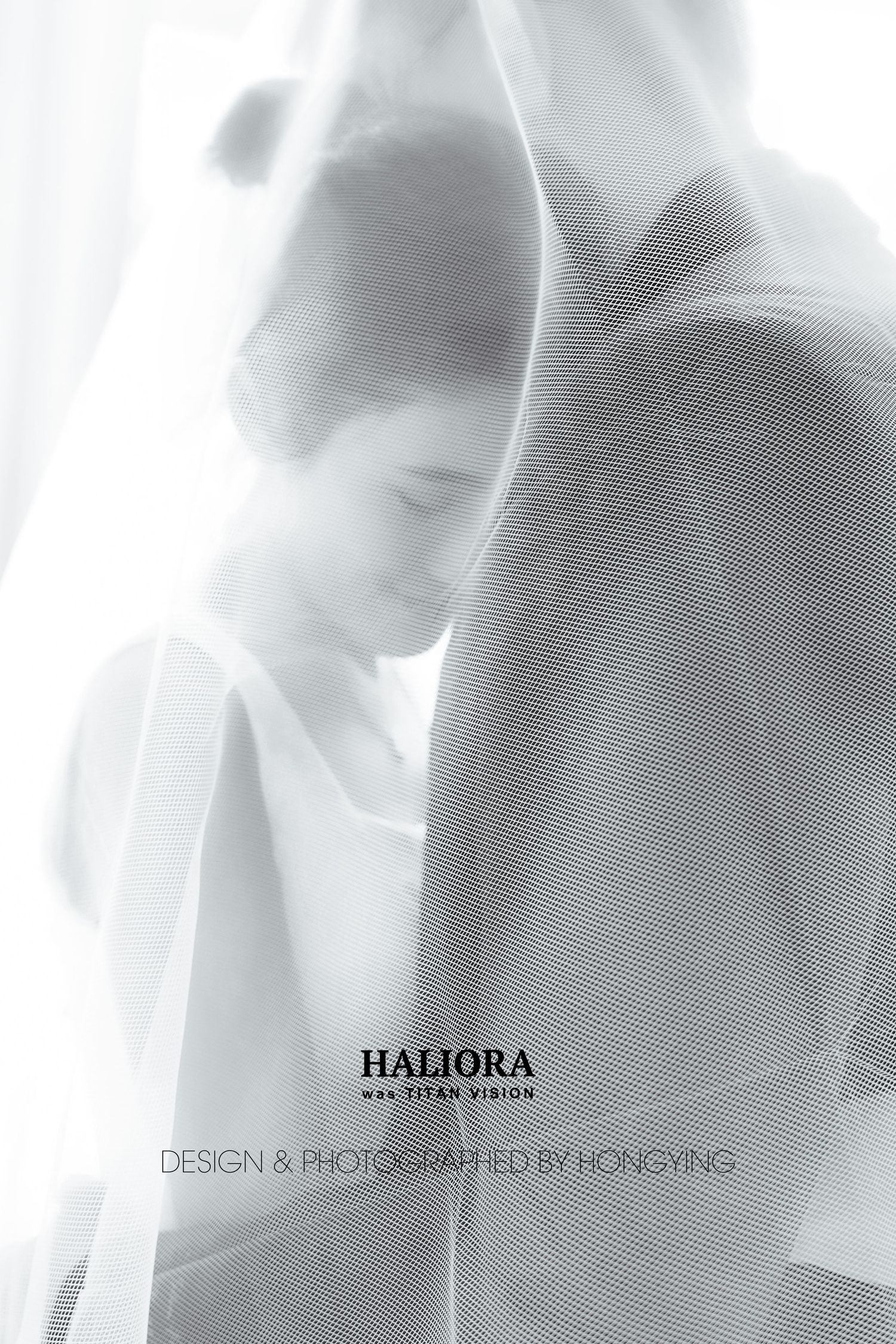 Artistic black and white photo of an Asian bride and groom partially veiled in soft white fabric near a window.