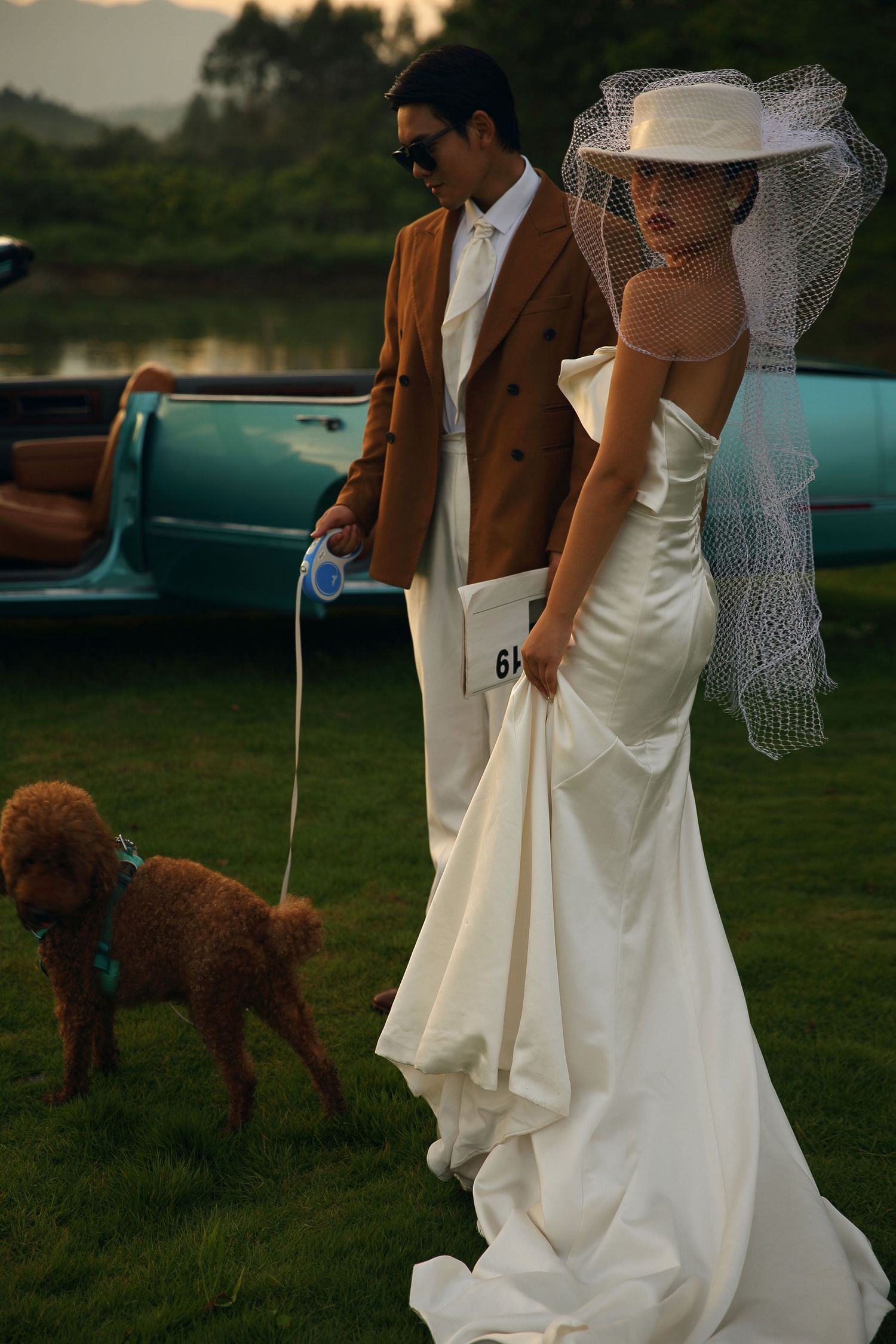 Bride in backless white gown and hat veil walks brown poodle with groom beside vintage teal car.