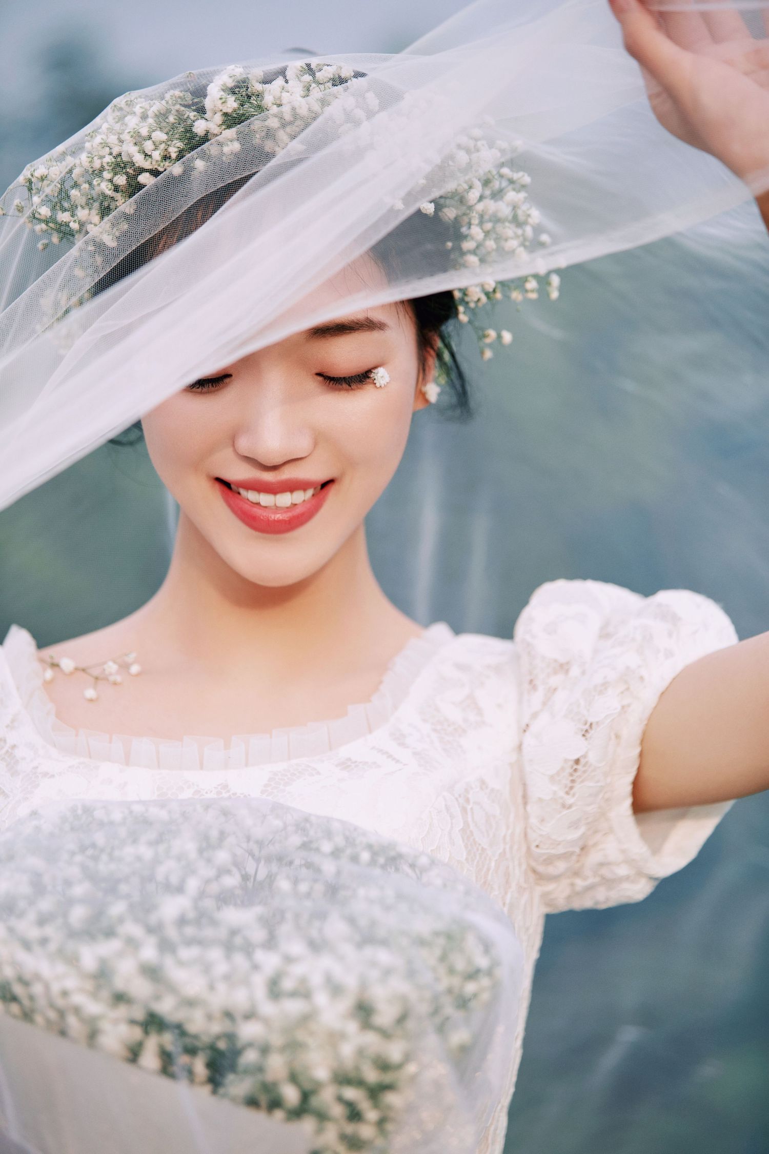 Radiant bride smiles softly under lifted veil, wearing floral crown and white lace dress with baby's breath.