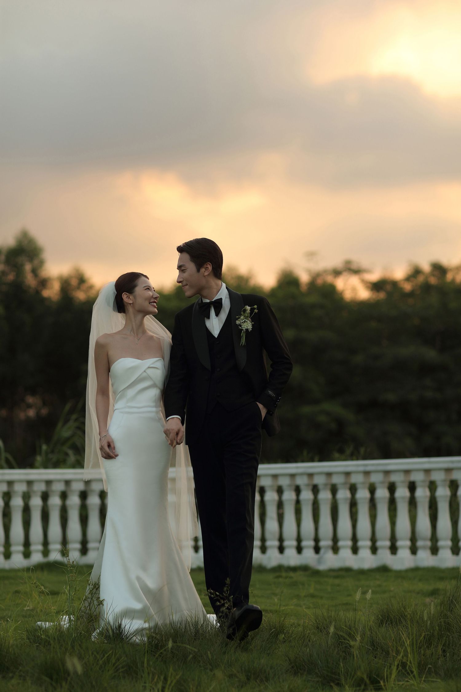 Bride in white strapless gown and groom in black tuxedo hold hands at sunset on lush green lawn.
