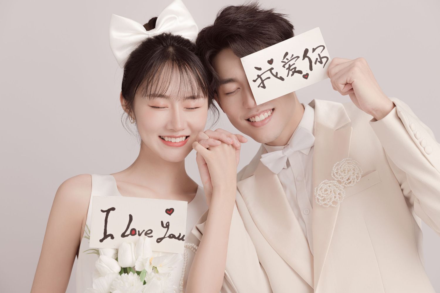Smiling couple in white wedding outfits posing closely, groom holding a love note card to his face.