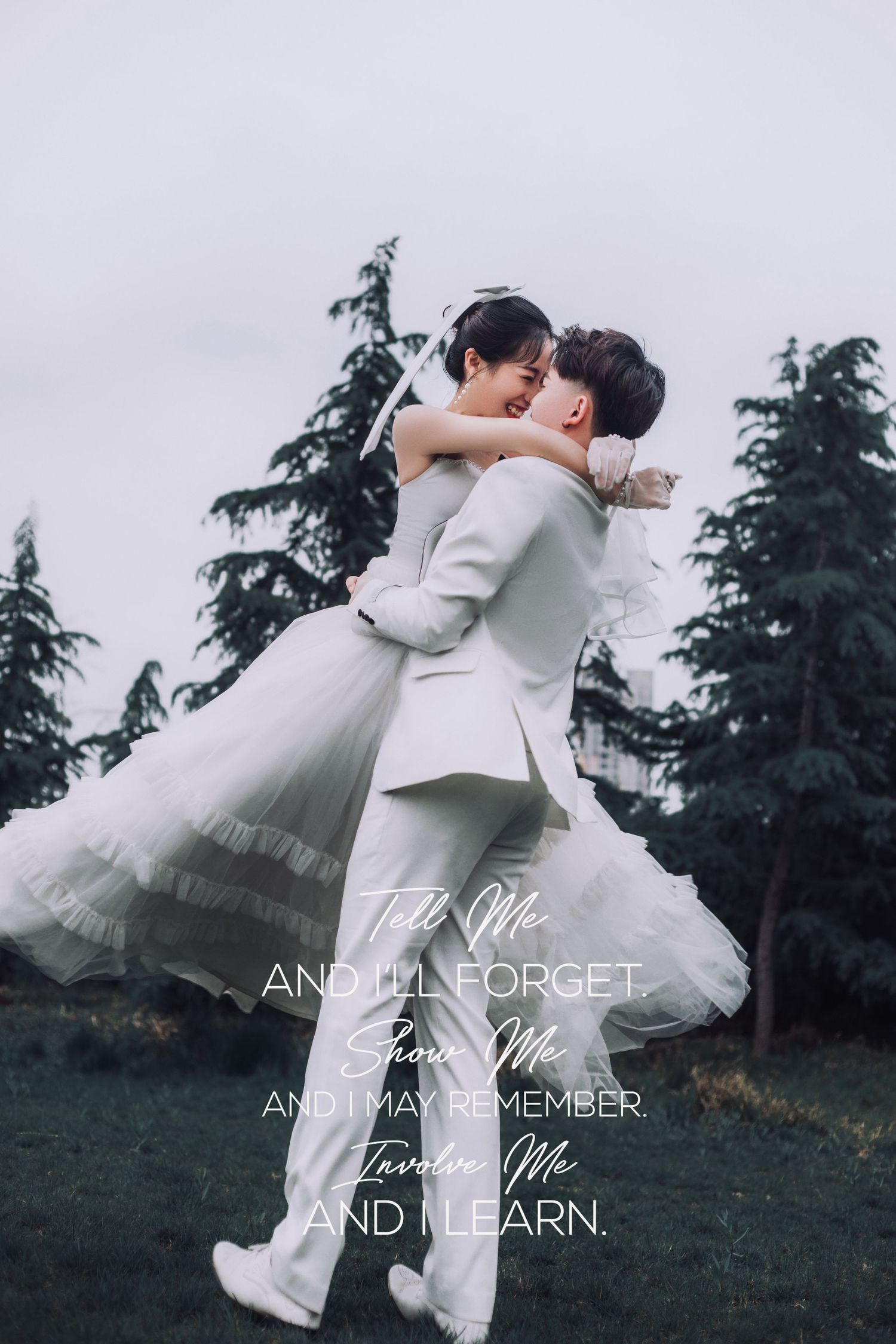 Happy couple embraces in wedding attire on lush green lawn with tall evergreen trees behind.