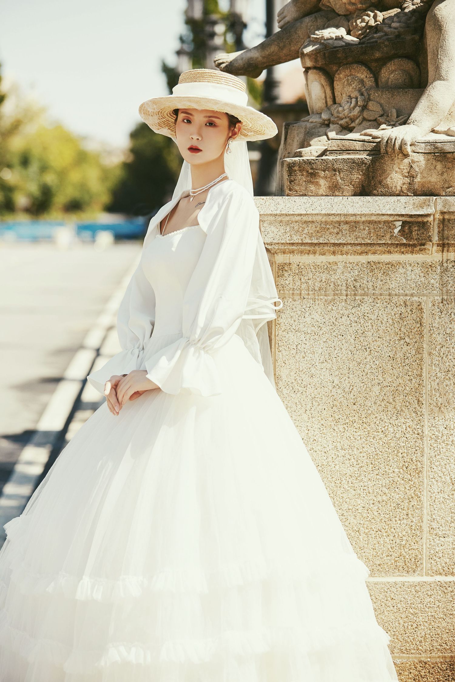 Elegant bride in white tulle gown and straw hat poses gracefully beside classical stone sculpture outdoors.