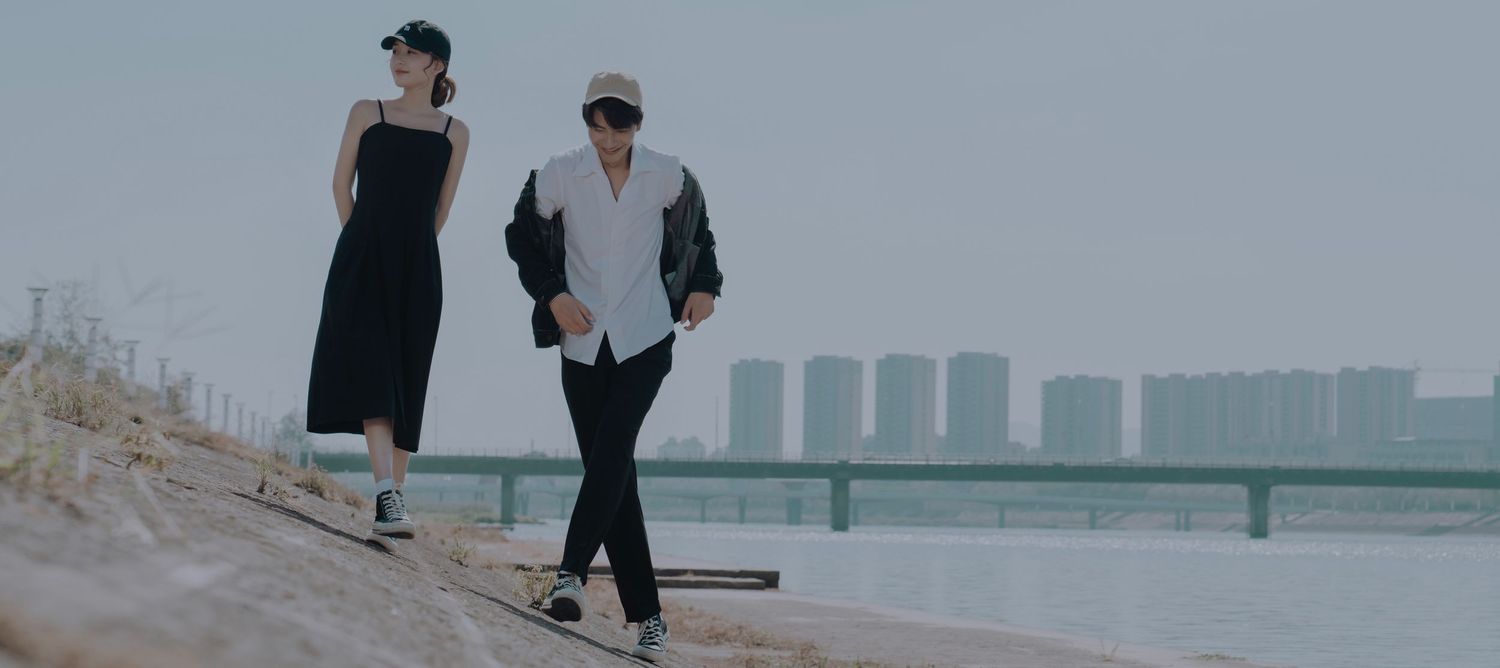 A young couple in black outfits walking along a scenic riverside path with a city skyline in the distance.