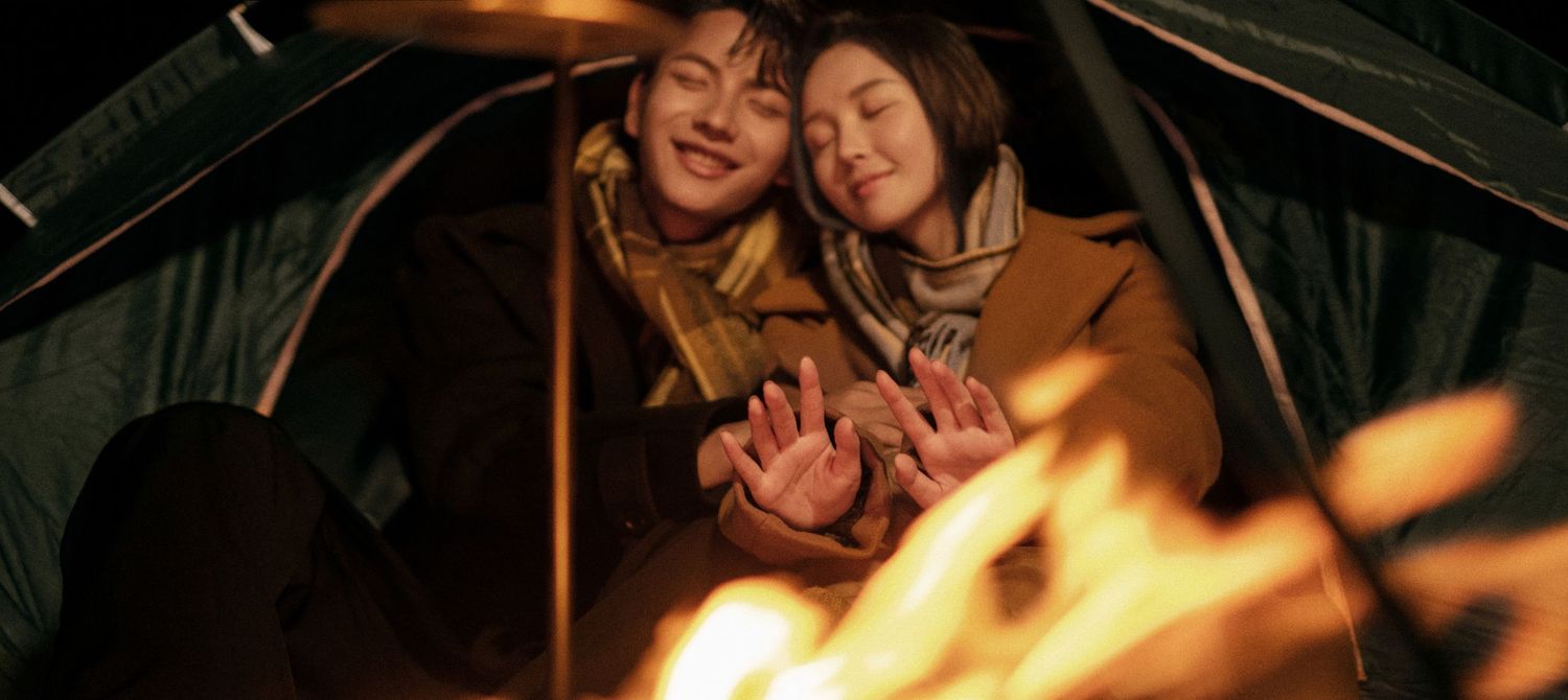 Two people smiling and warming hands by a glowing campfire inside a tent.