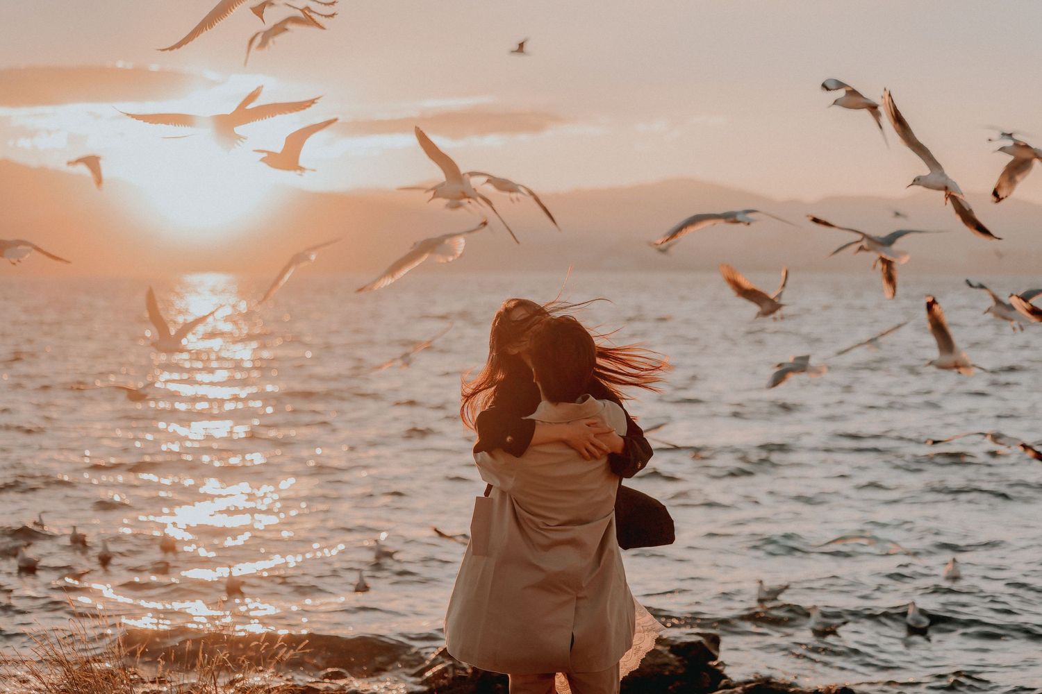 A couple embraces on rocks by the ocean at golden sunset with seagulls flying overhead.