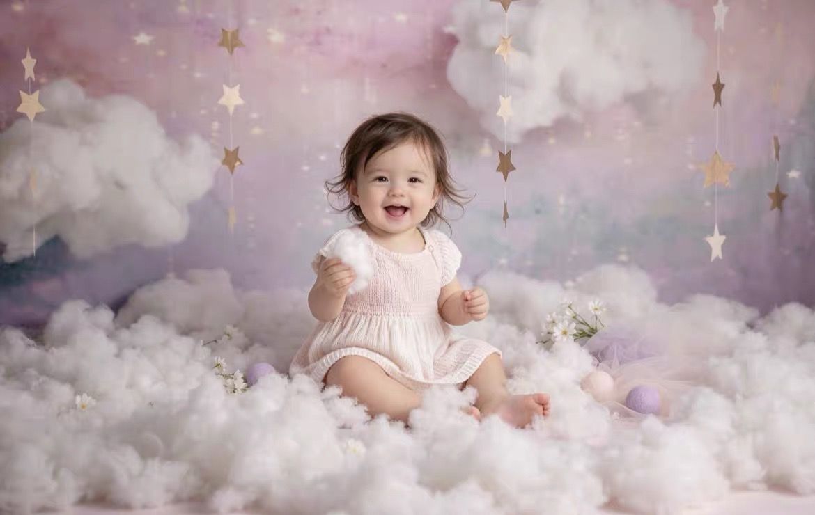 Smiling baby girl in white dress sitting among fluffy clouds with stars on a dreamy pink purple backdrop.