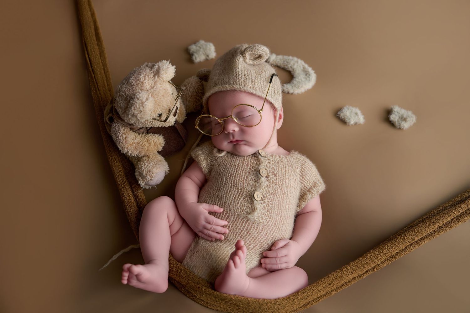 Sleeping newborn in knit bear outfit and glasses posed in hammock with teddy bear on brown background.