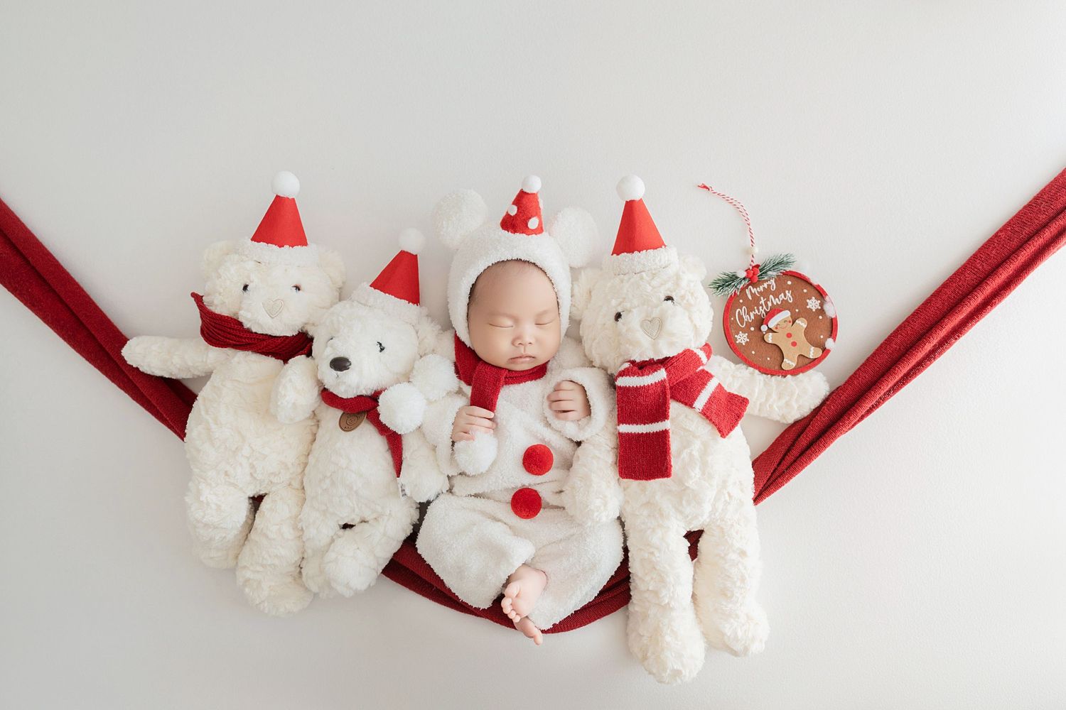 Sleeping newborn in Santa outfit lying in red hammock flanked by white Christmas teddy bears on white background.