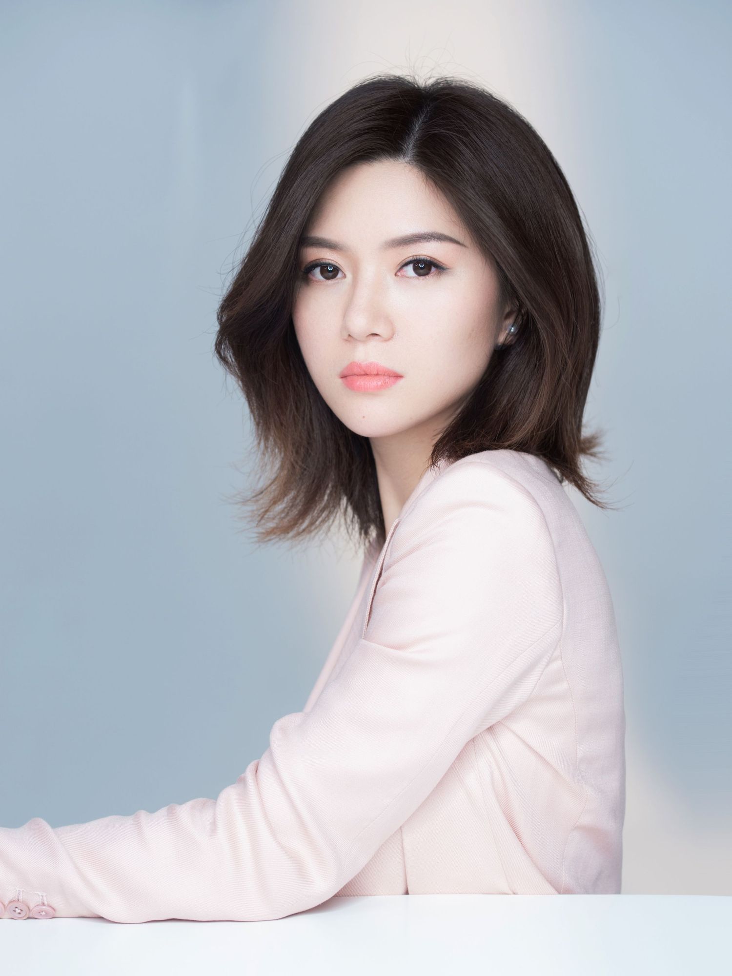 Elegant Asian woman with dark bob haircut in a pink long-sleeve top posing against a soft blue background.