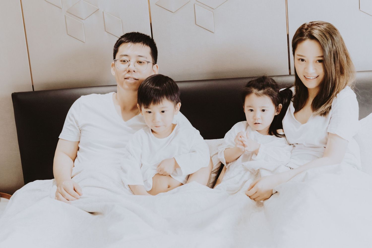 An Asian family of four sits together on a white bed, parents and two toddlers posing for a portrait.