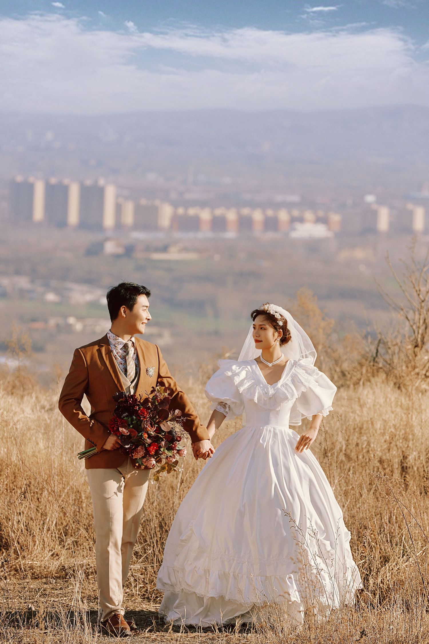 Couple in vintage-style wedding attire walk through golden autumn field with city skyline backdrop.