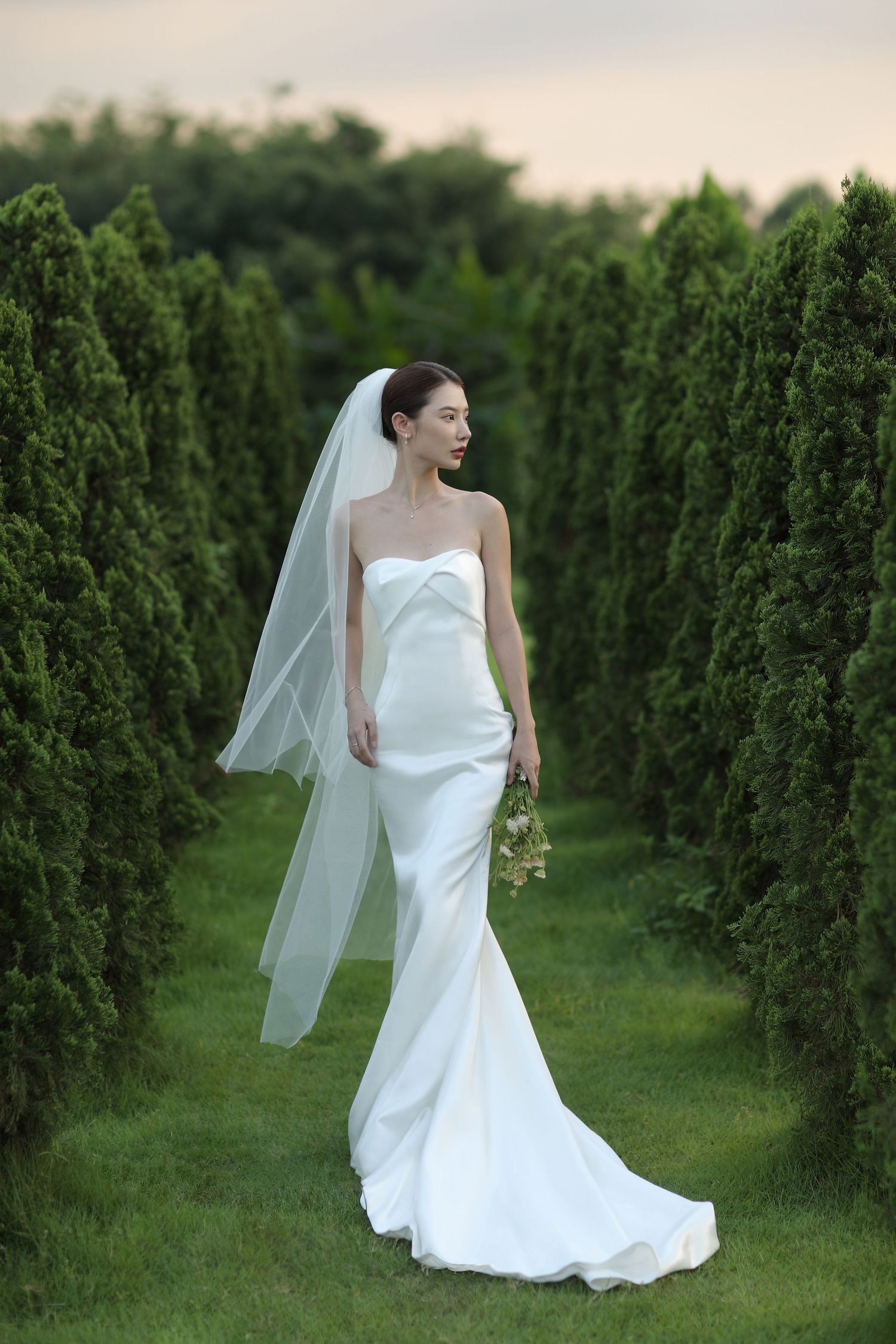 Bride in a sleek white strapless gown and veil stands gracefully between rows of tall green cypress trees.
