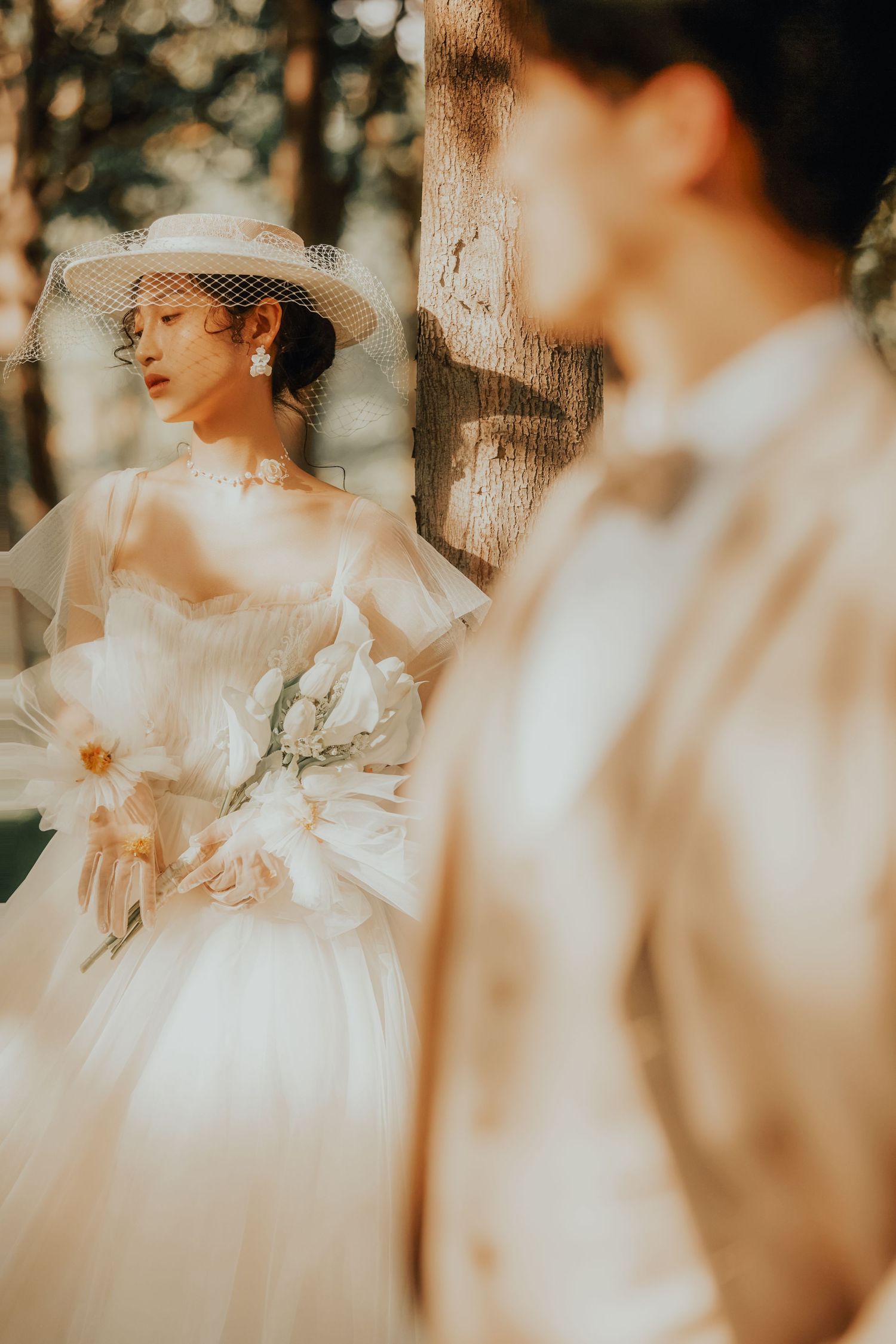 Elegant Asian bride in a white ballgown and veiled hat holds a floral bouquet beside a tree in warm sunlight.