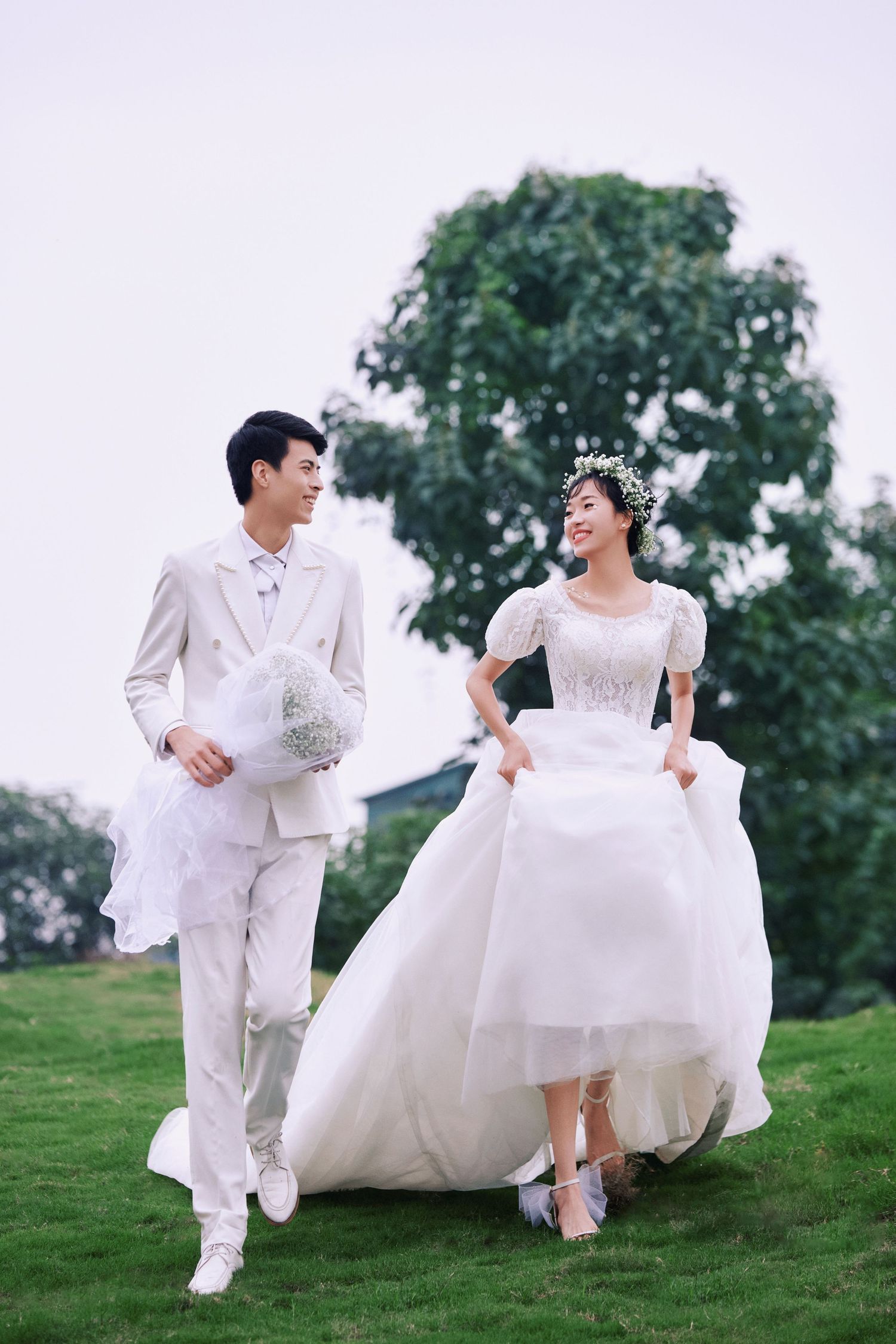 Happy wedding couple walking hand in hand on green lawn, bride lifting gown, groom holding bouquet.