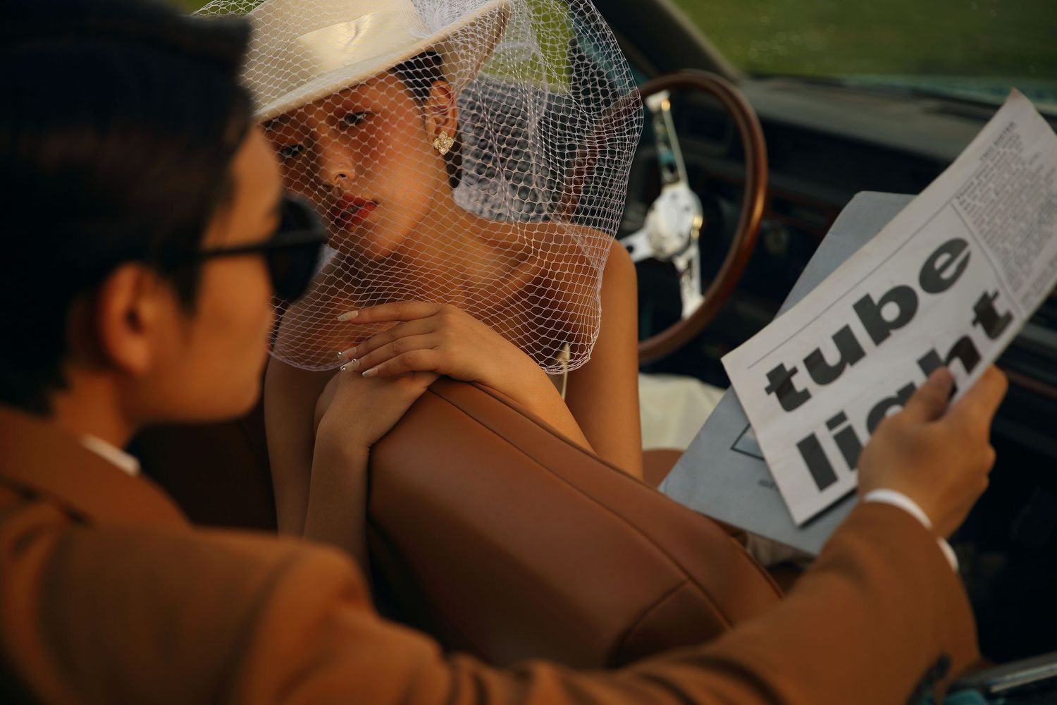 Stylish bride in white hat veil reads magazine beside groom in vintage classic car interior.