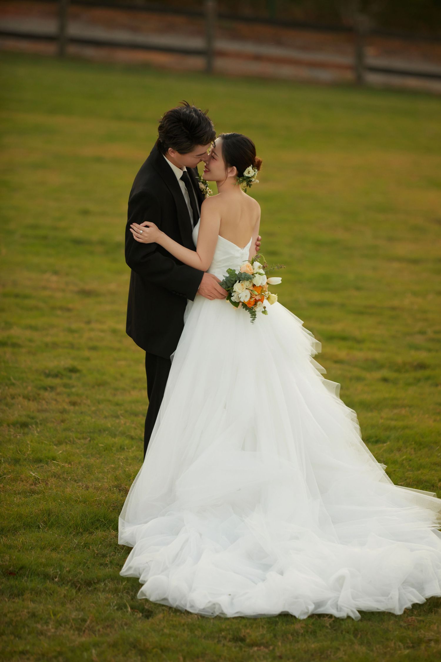 Bride and groom share romantic kiss on green lawn, bride holding colorful bouquet in white ballgown.