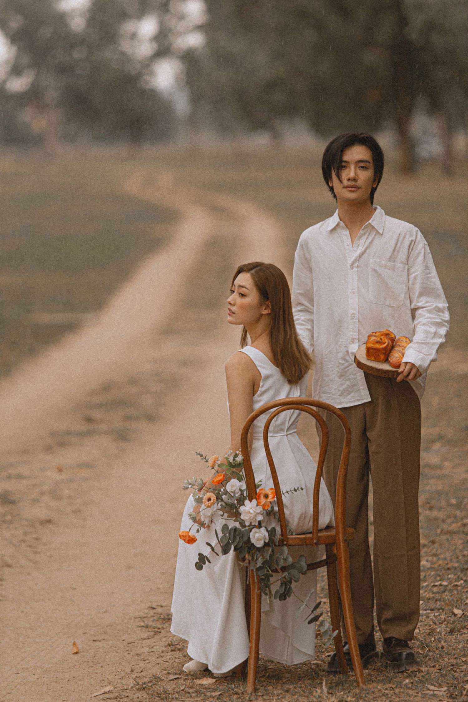 Asian couple in white and tan outfits pose on a dirt road with flowers and a wooden chair in golden light.
