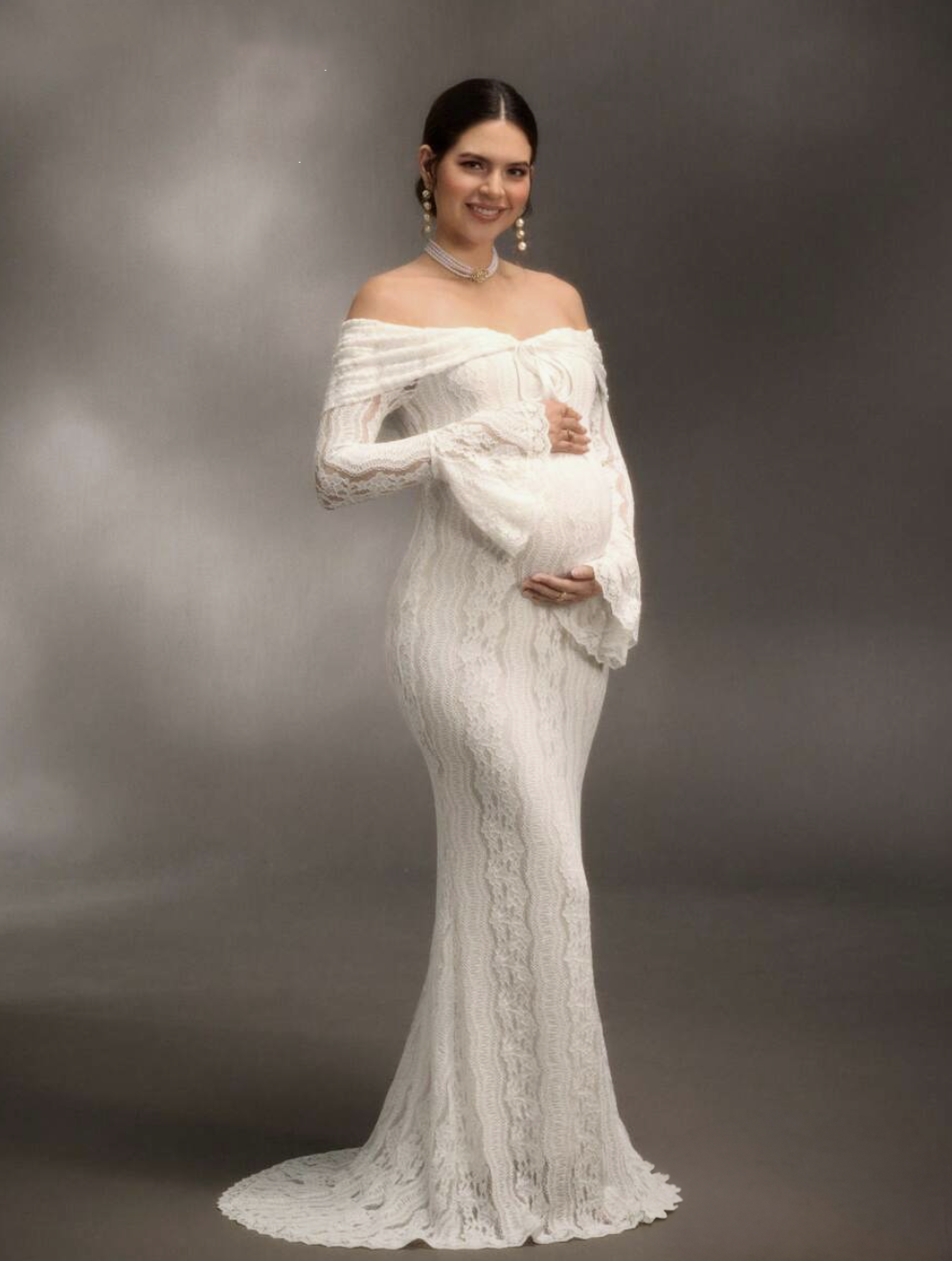 Pregnant woman in elegant white off-shoulder lace maternity gown with long sleeves and train.
