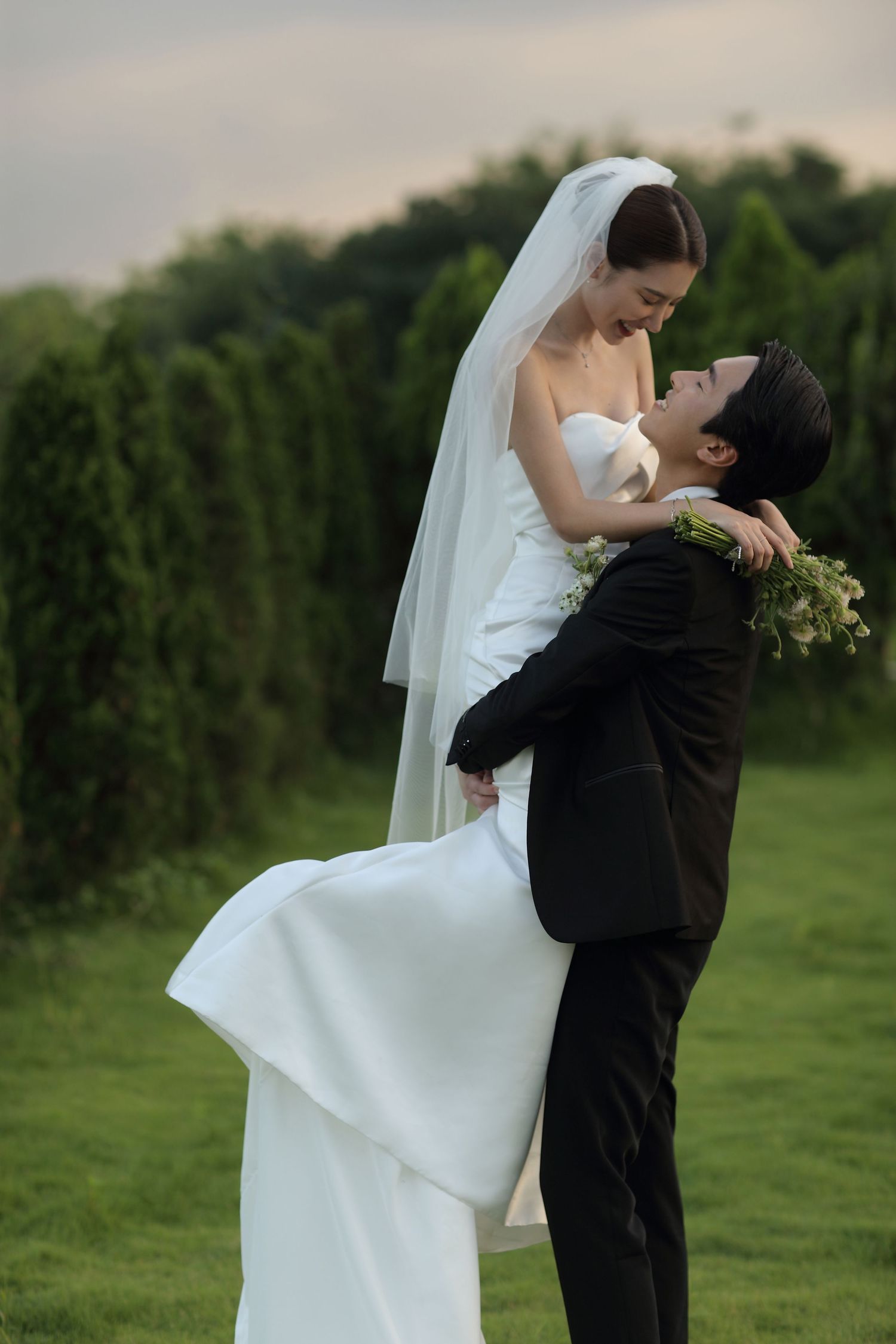Groom in black suit lifts smiling bride in white wedding dress and veil in a garden setting.