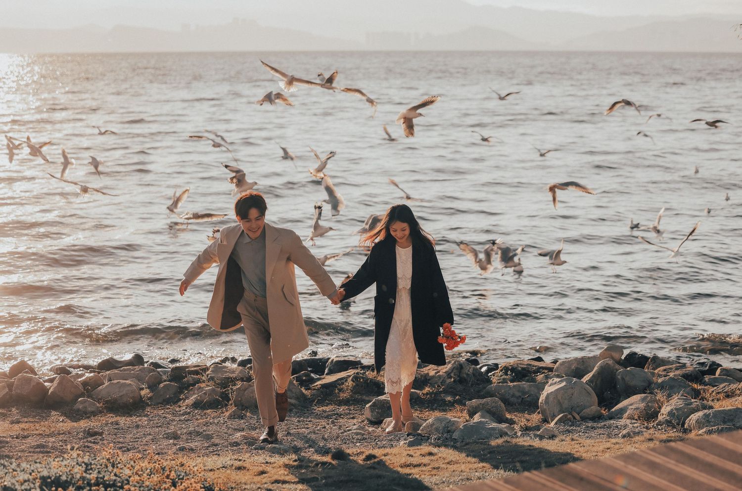 A couple walks hand in hand along a rocky shoreline with seagulls soaring above the water.