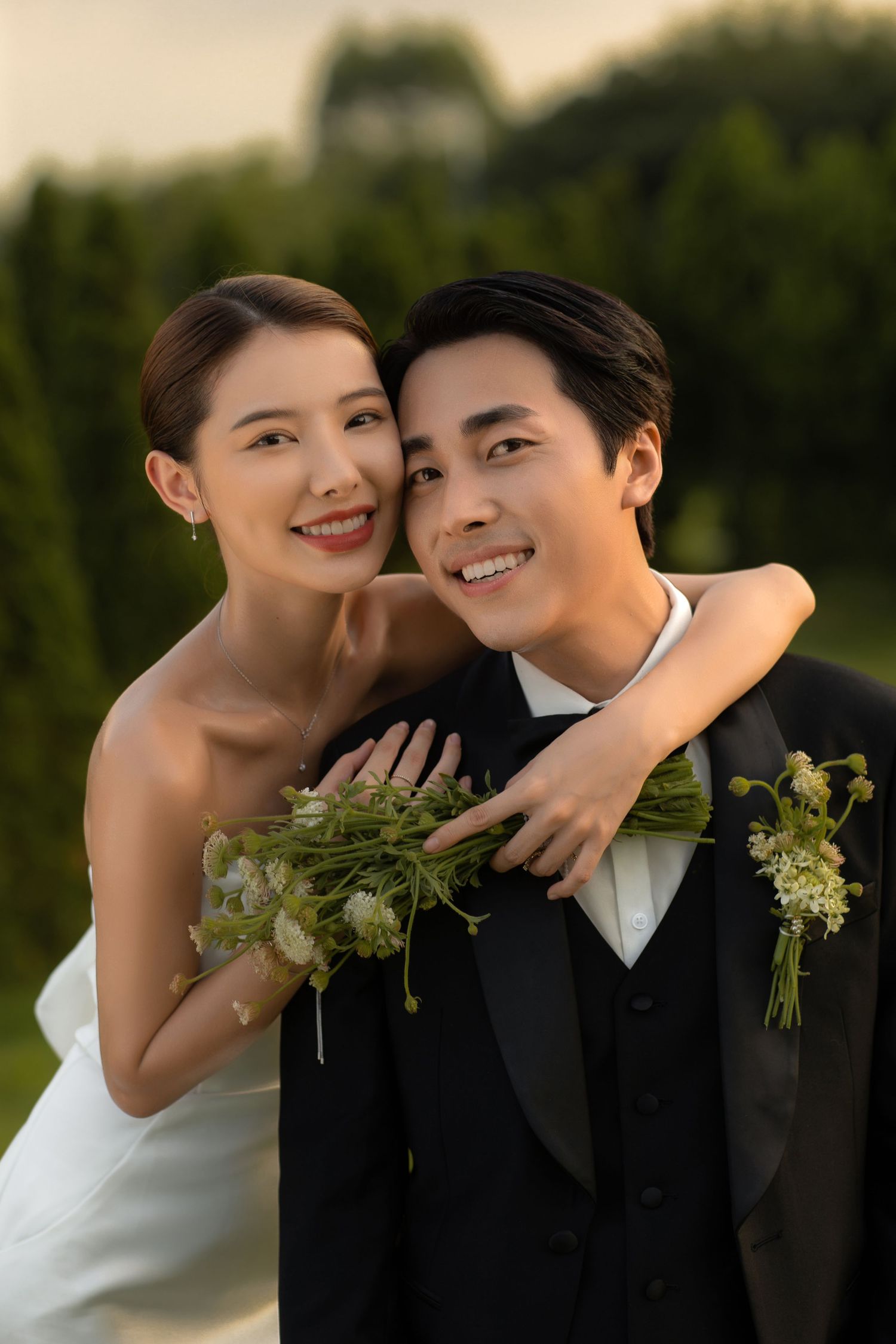 Smiling bride and groom pose outdoors, she holds a bouquet and wraps her arms around him from behind.