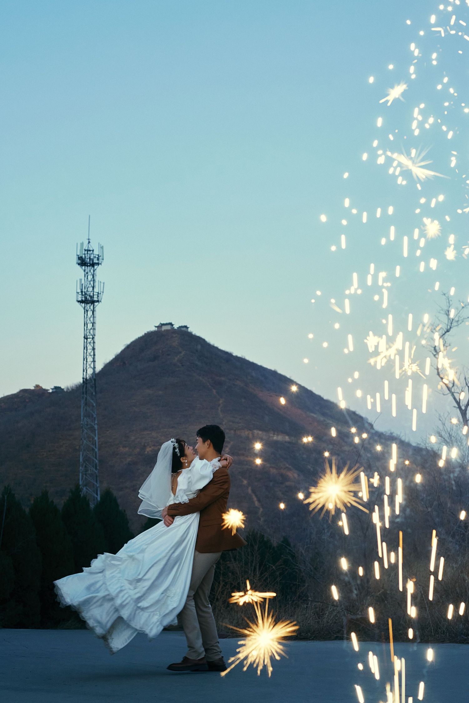Groom lifts bride in white wedding dress amid sparkling fireworks with mountain backdrop at dusk.