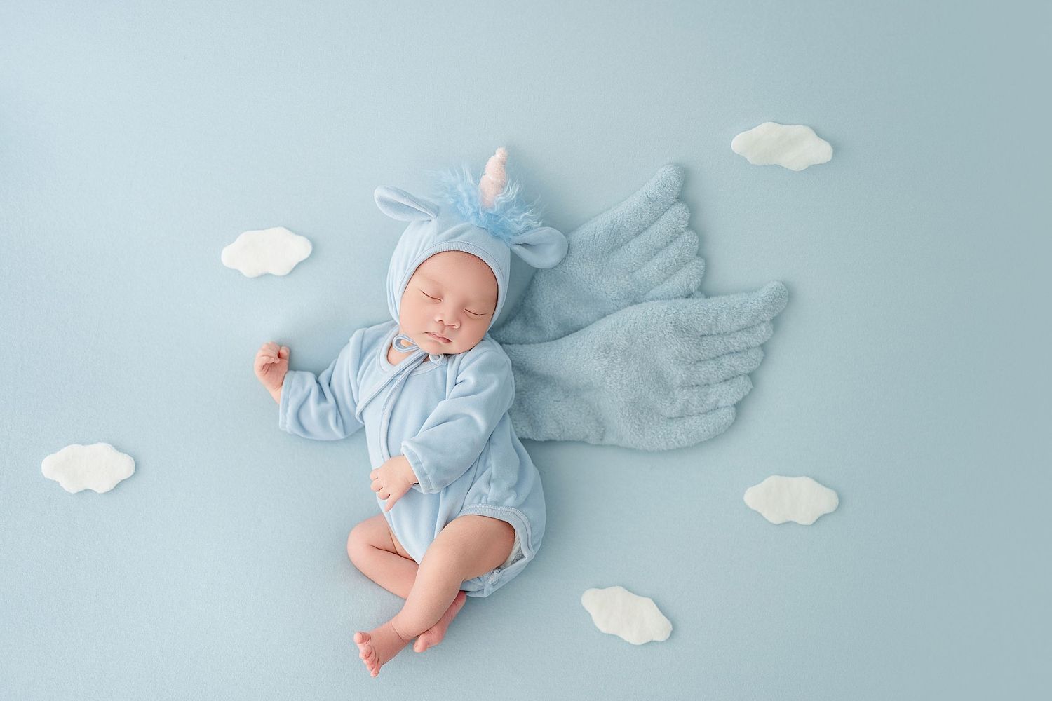 Newborn baby in blue angel outfit with wings and cloud props on light blue background.