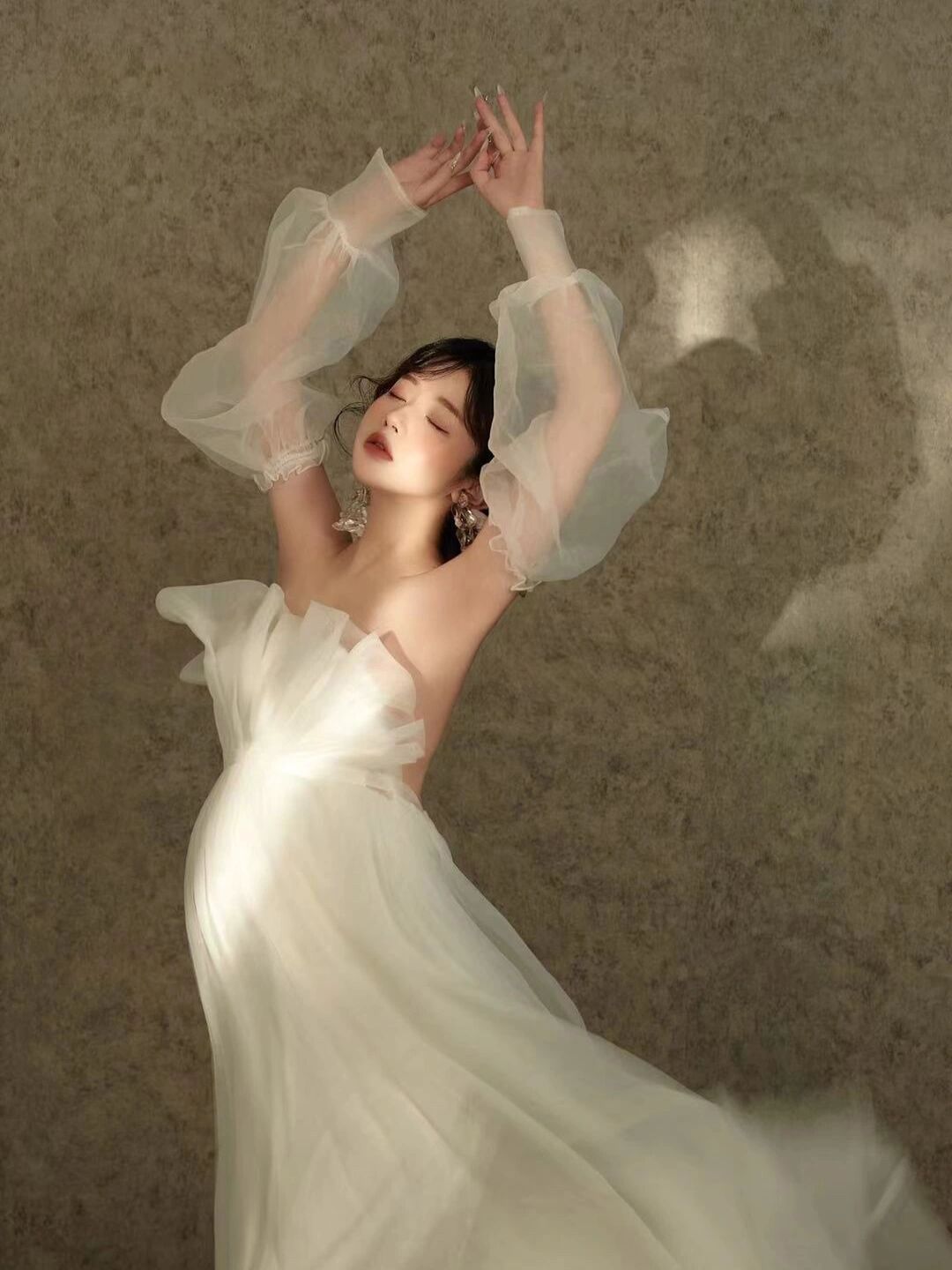 Elegant woman in flowing white tulle gown with sheer puff sleeves poses gracefully against neutral backdrop.