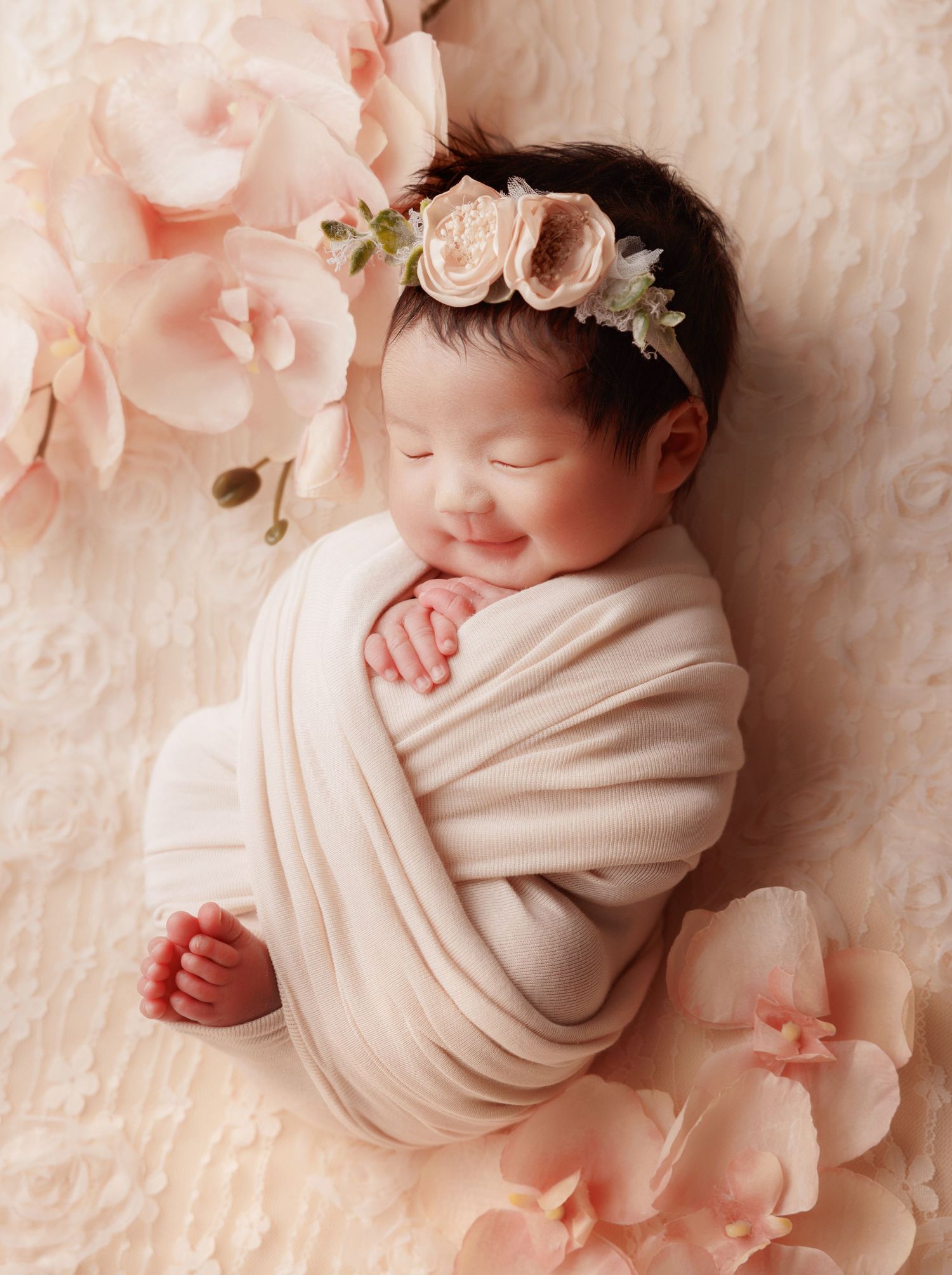 Smiling swaddled newborn baby girl with pink floral headband among pink blooms.