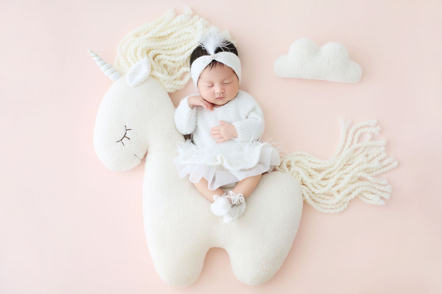 Newborn baby in white outfit with panda hat sleeping on a unicorn plush toy on a soft pink background.