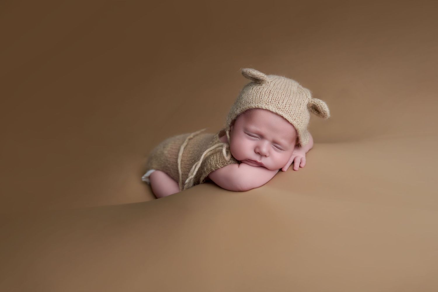 Sleeping newborn baby in knit bear bonnet and romper posed on tan background.