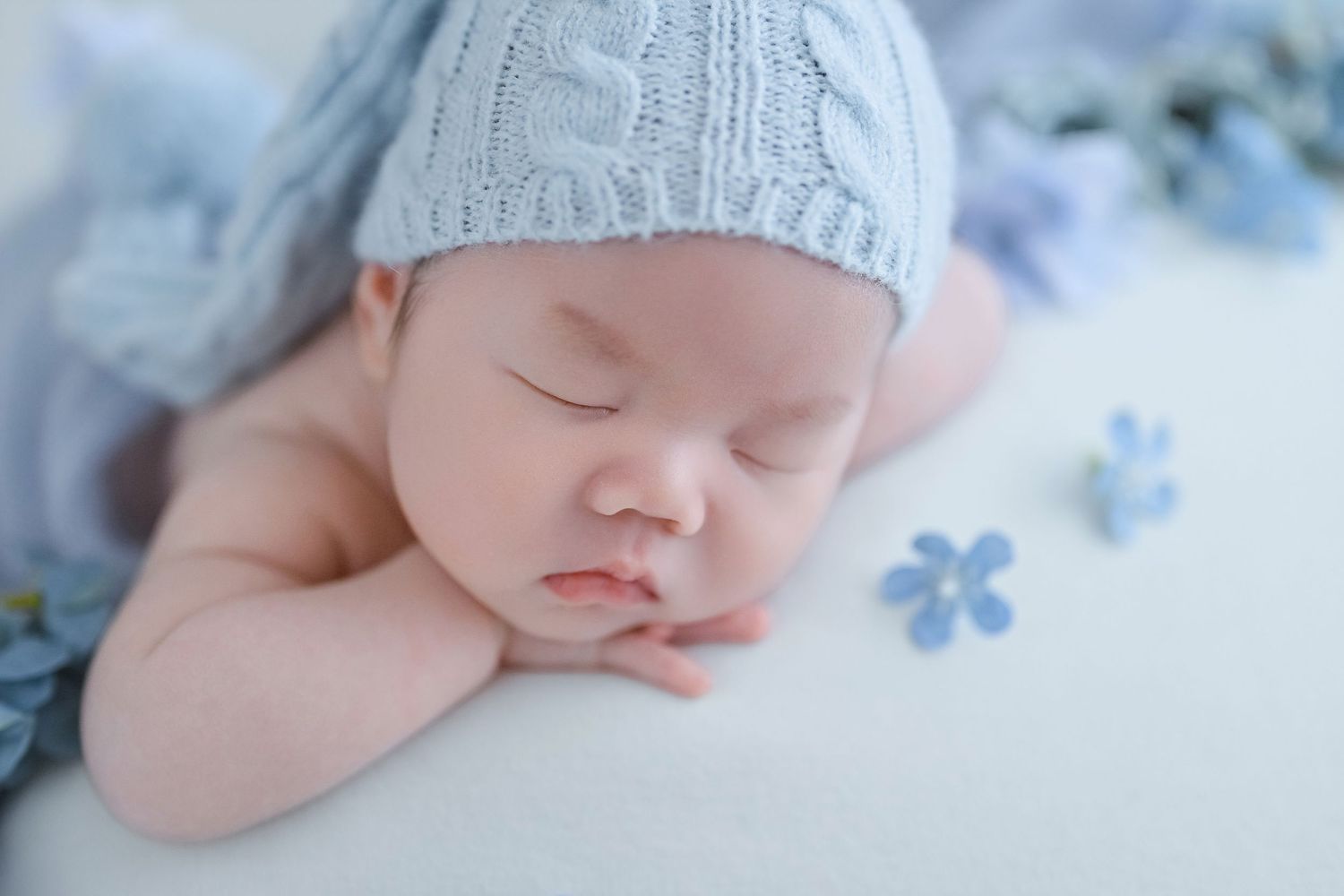 Close-up of sleeping newborn in blue knit hat with small blue flowers on white background.
