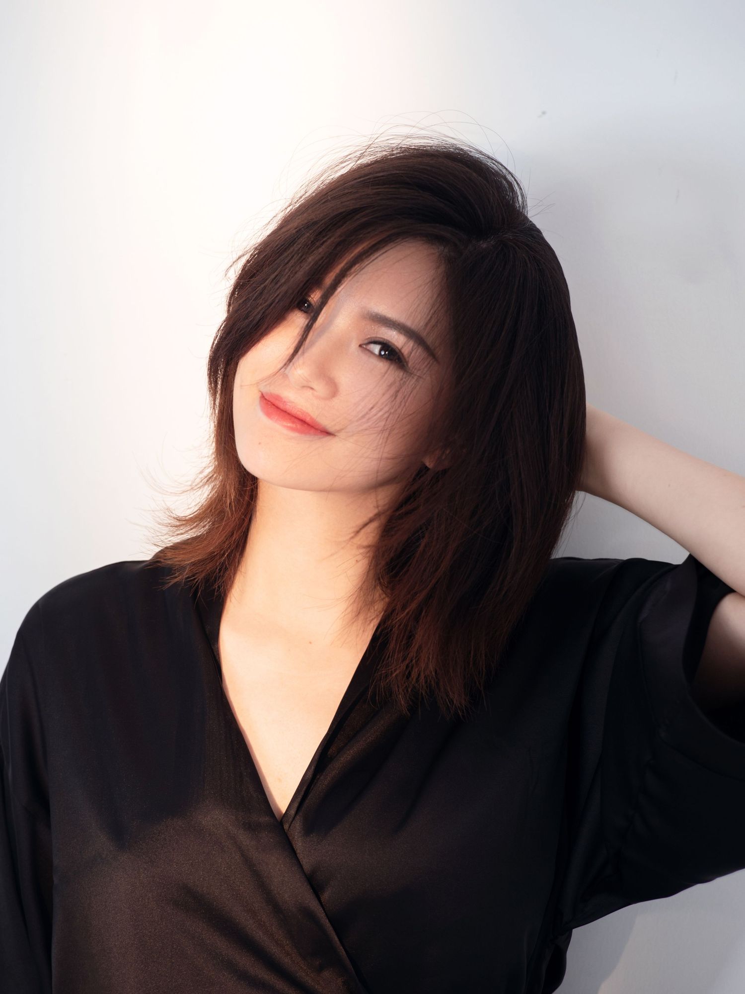 Young Asian woman with shoulder-length dark hair wearing a black wrap top, smiling against a white wall.