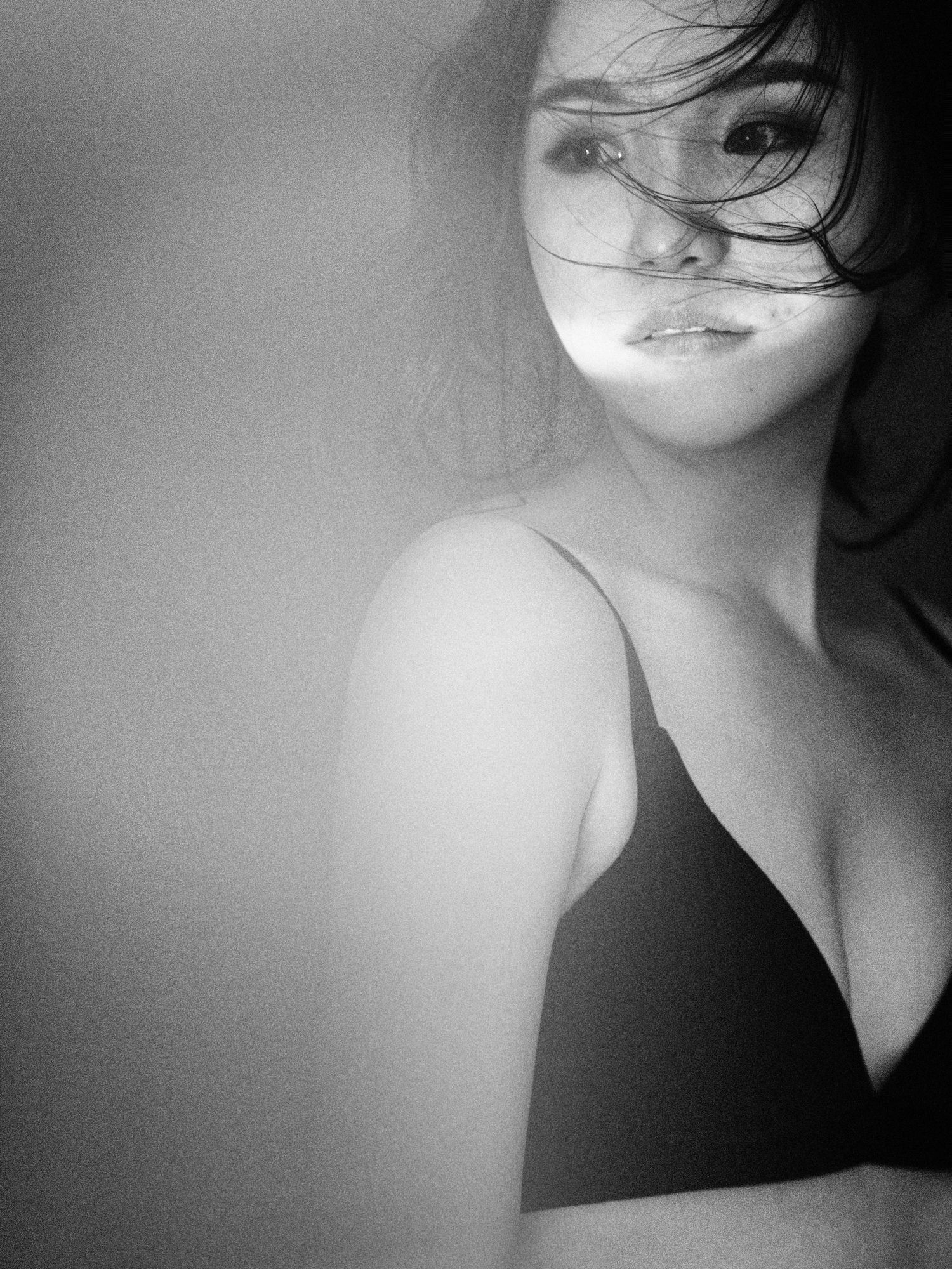 Black and white portrait of a young Asian woman in a black bra with windswept hair gazing softly at the camera.