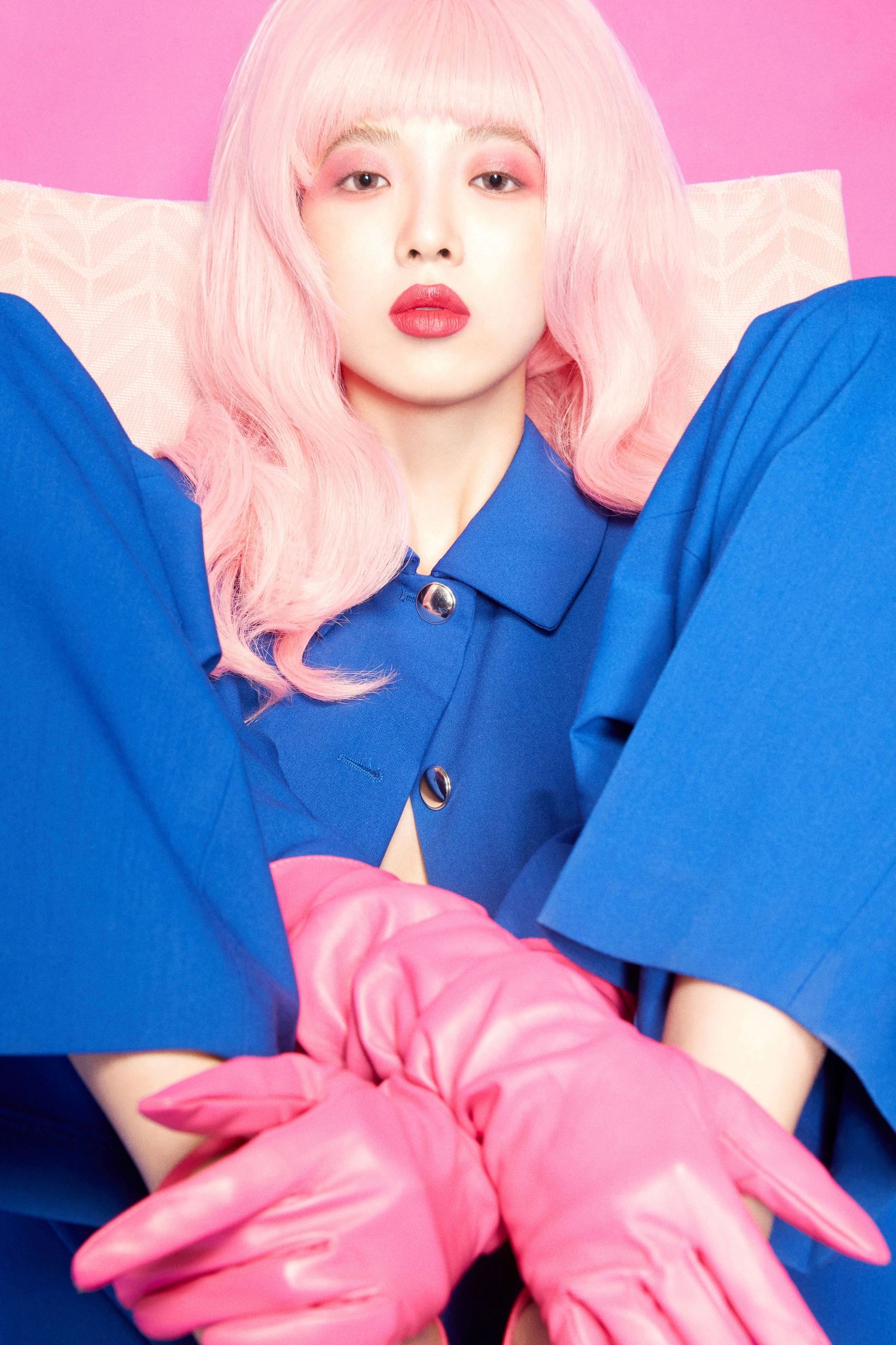 Woman with pink hair wearing blue coat and pink leather gloves poses against a vibrant pink background.