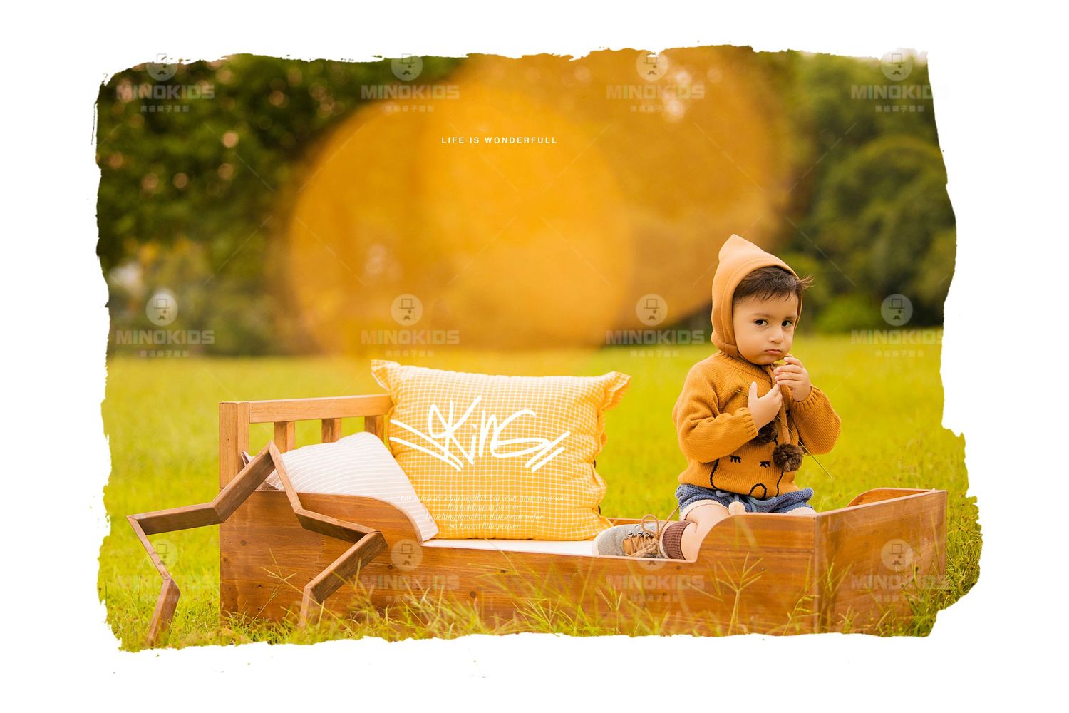 Toddler in yellow hooded jacket sits on wooden bed prop in sunlit green grass with lens flare.