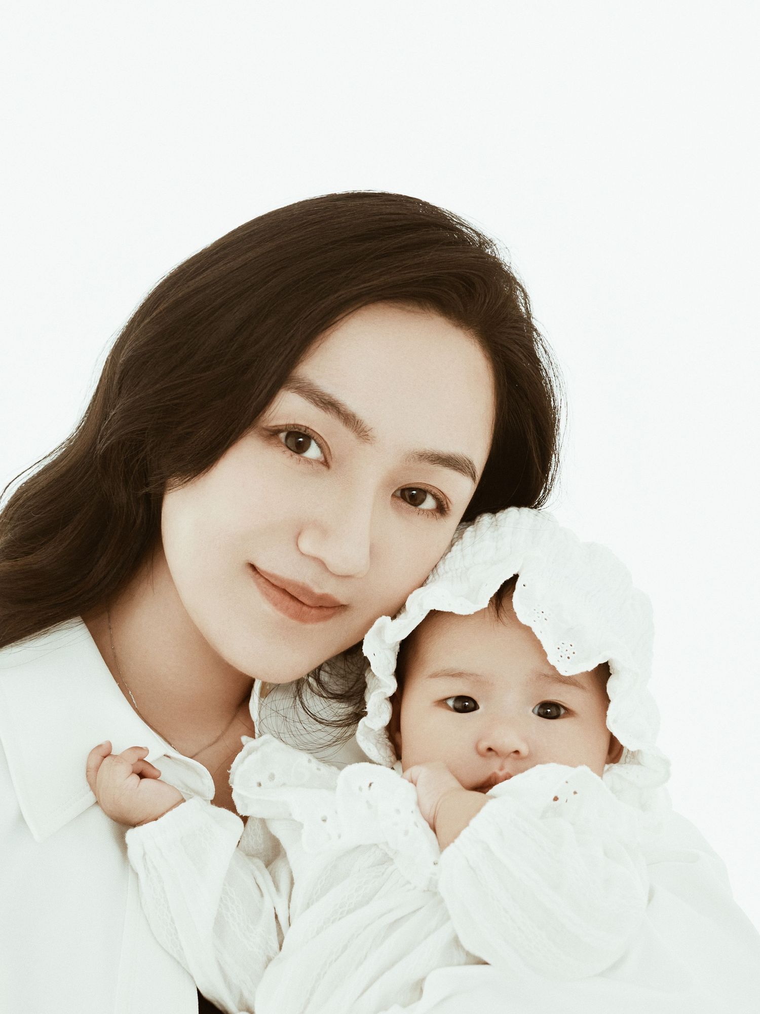 Beautiful Asian mother with long dark hair poses with her adorable newborn baby in white frilly outfit.