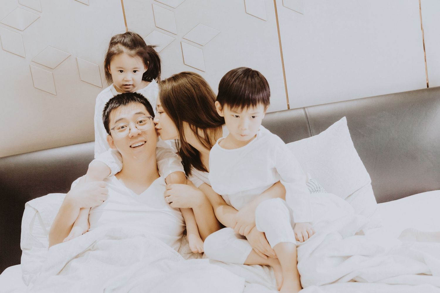 A happy Asian family cuddles together on a white bed, with two young children embracing their parents warmly.