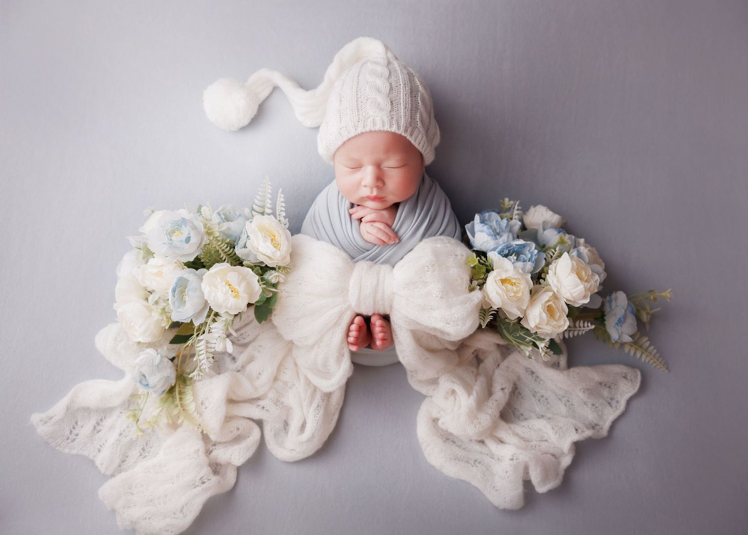 Sleeping newborn baby in white knit hat posed with large bow and blue and white floral bouquets on gray backdrop.