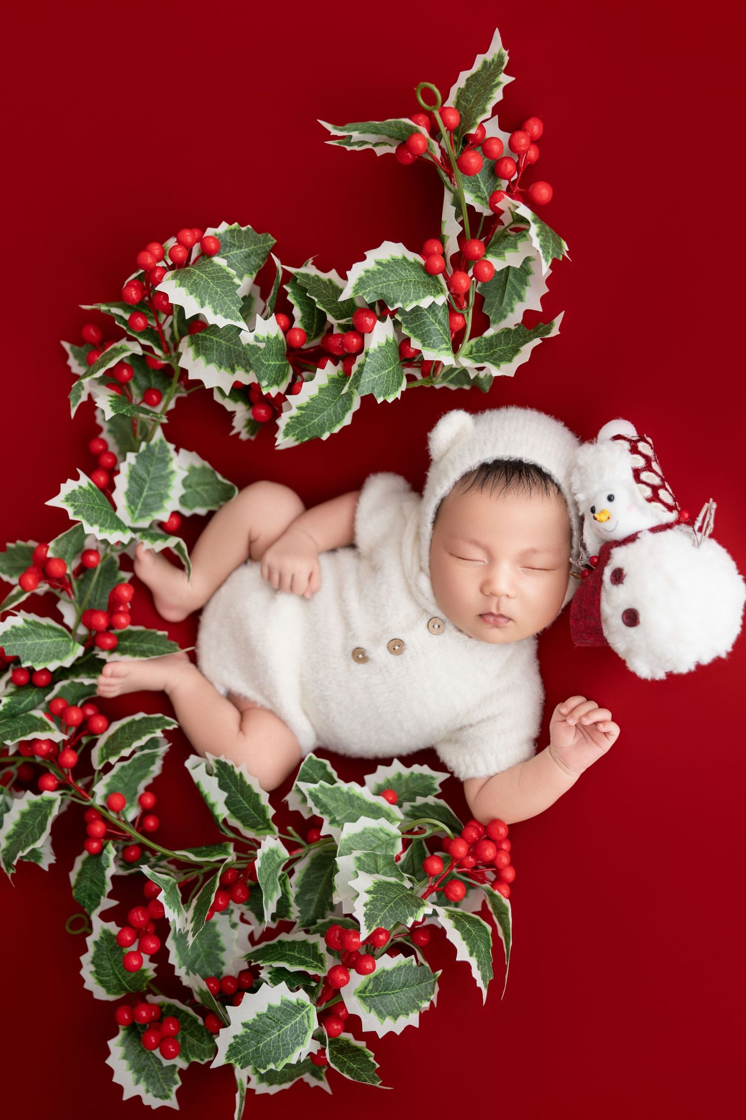 Peaceful newborn baby in white bear outfit sleeping in holly wreath with red berries and snowman toy on red background.
