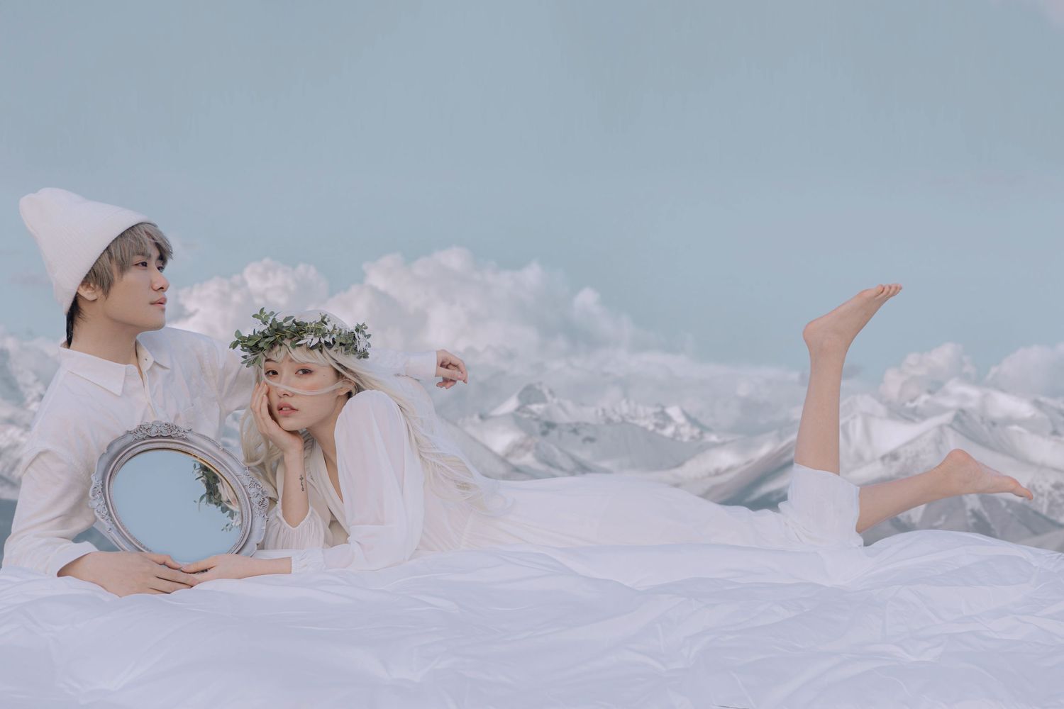 Couple in white outfits lying on white surface, man holding round mirror, woman wearing floral crown.