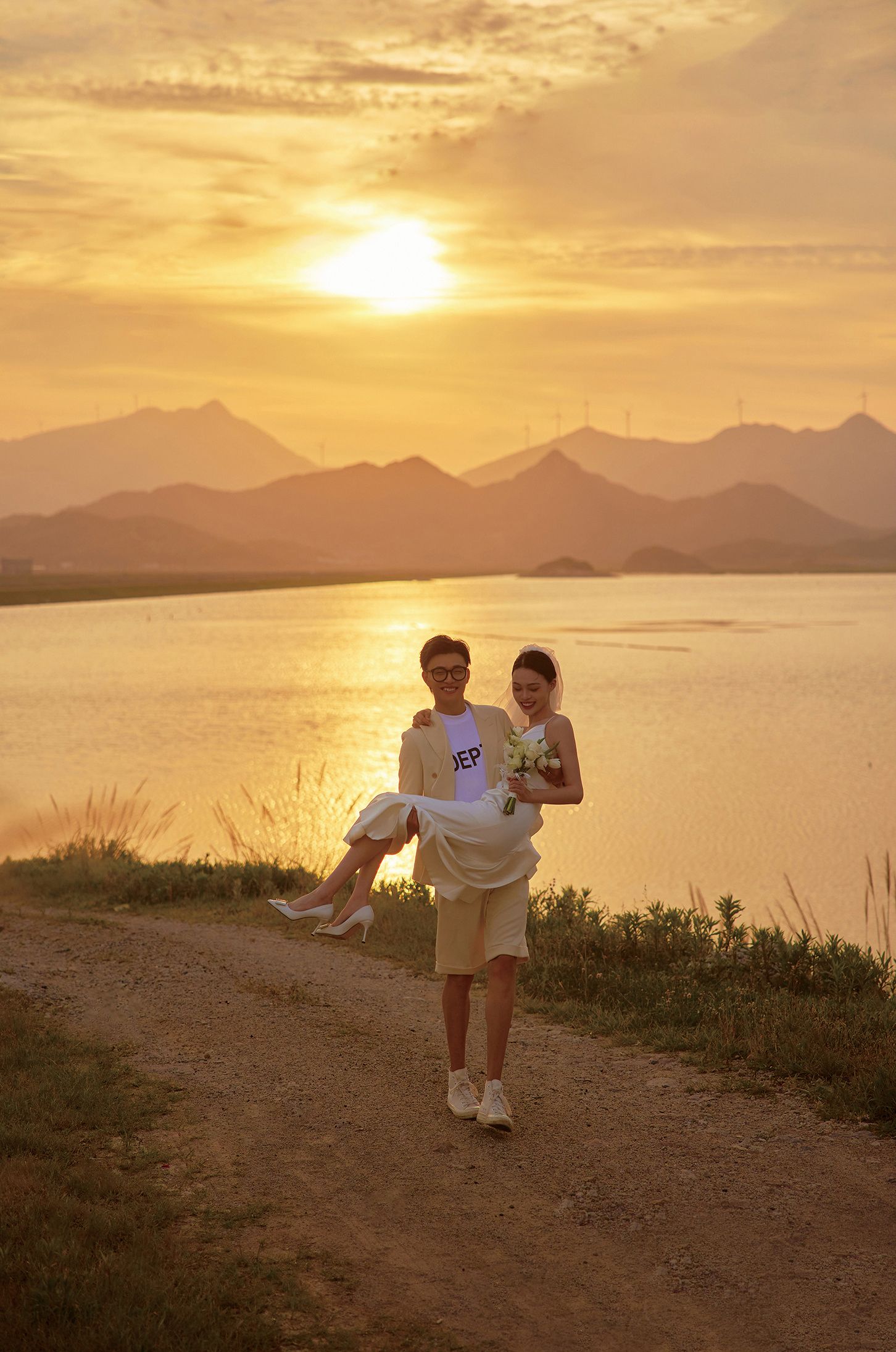 Groom carrying bride in his arms on a dirt path with a golden sunset and mountain lake backdrop.
