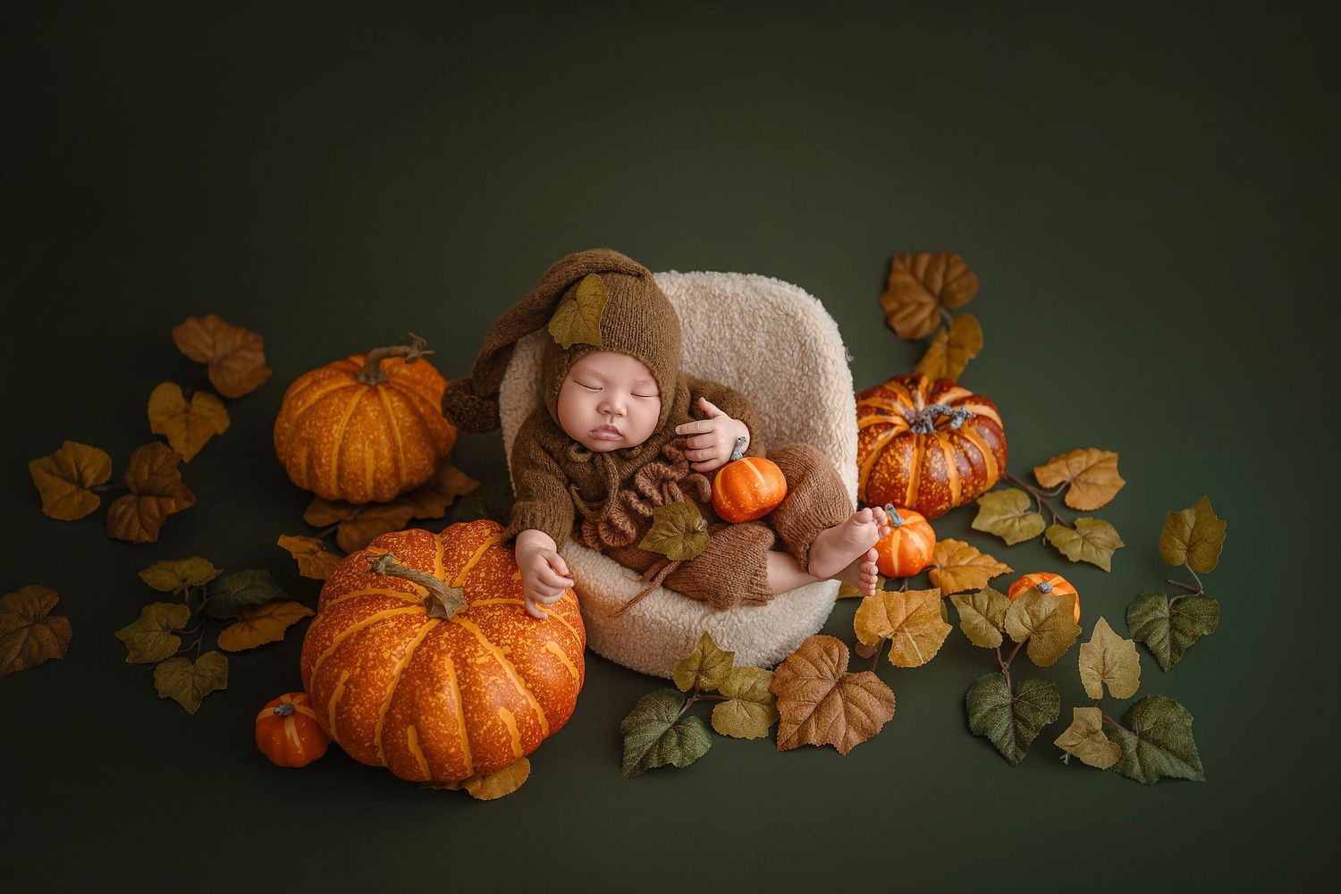 Newborn baby in brown knit hat and romper sleeps in a chair surrounded by orange pumpkins and autumn leaves.