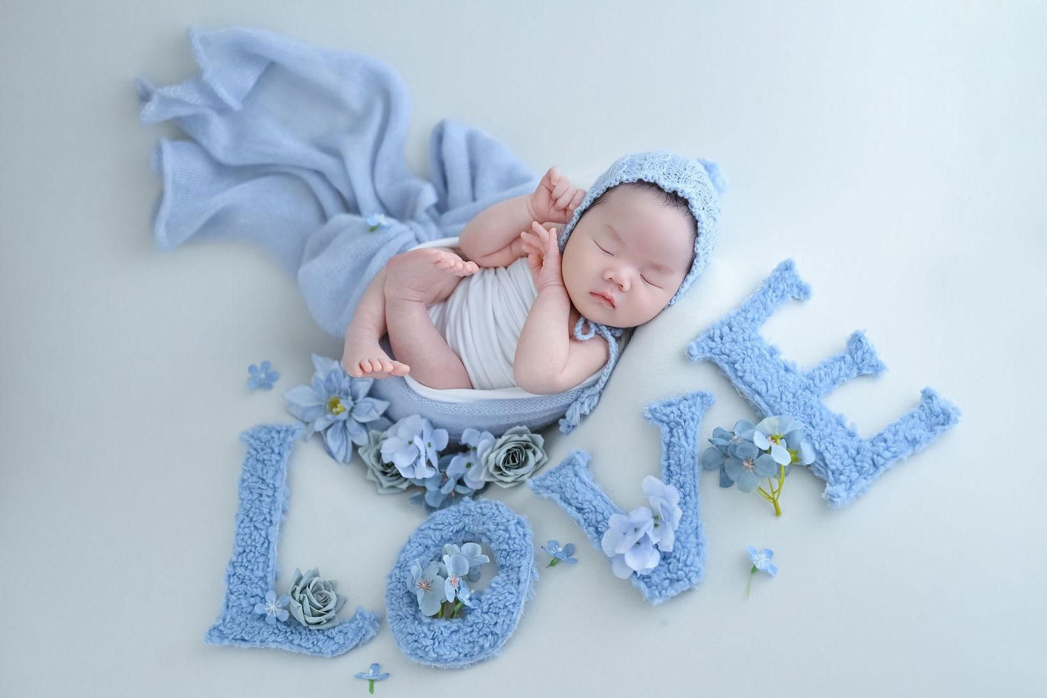 Sleeping newborn in blue bonnet posed in bowl beside knit LOVE letters and blue flowers.