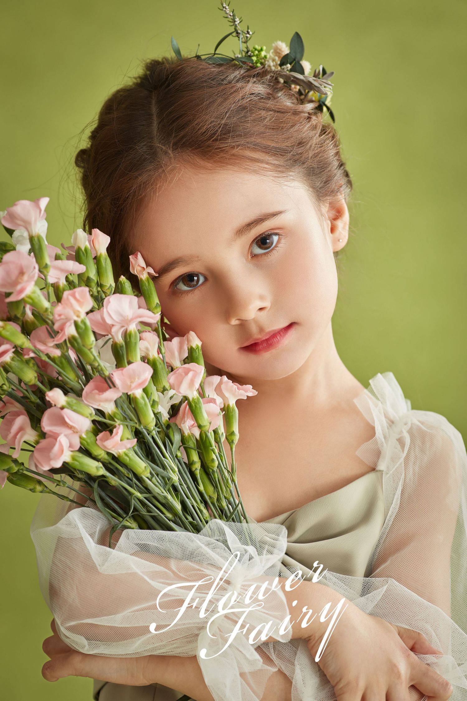 Young girl in white dress holds pink flowers bouquet wearing floral crown against green background.